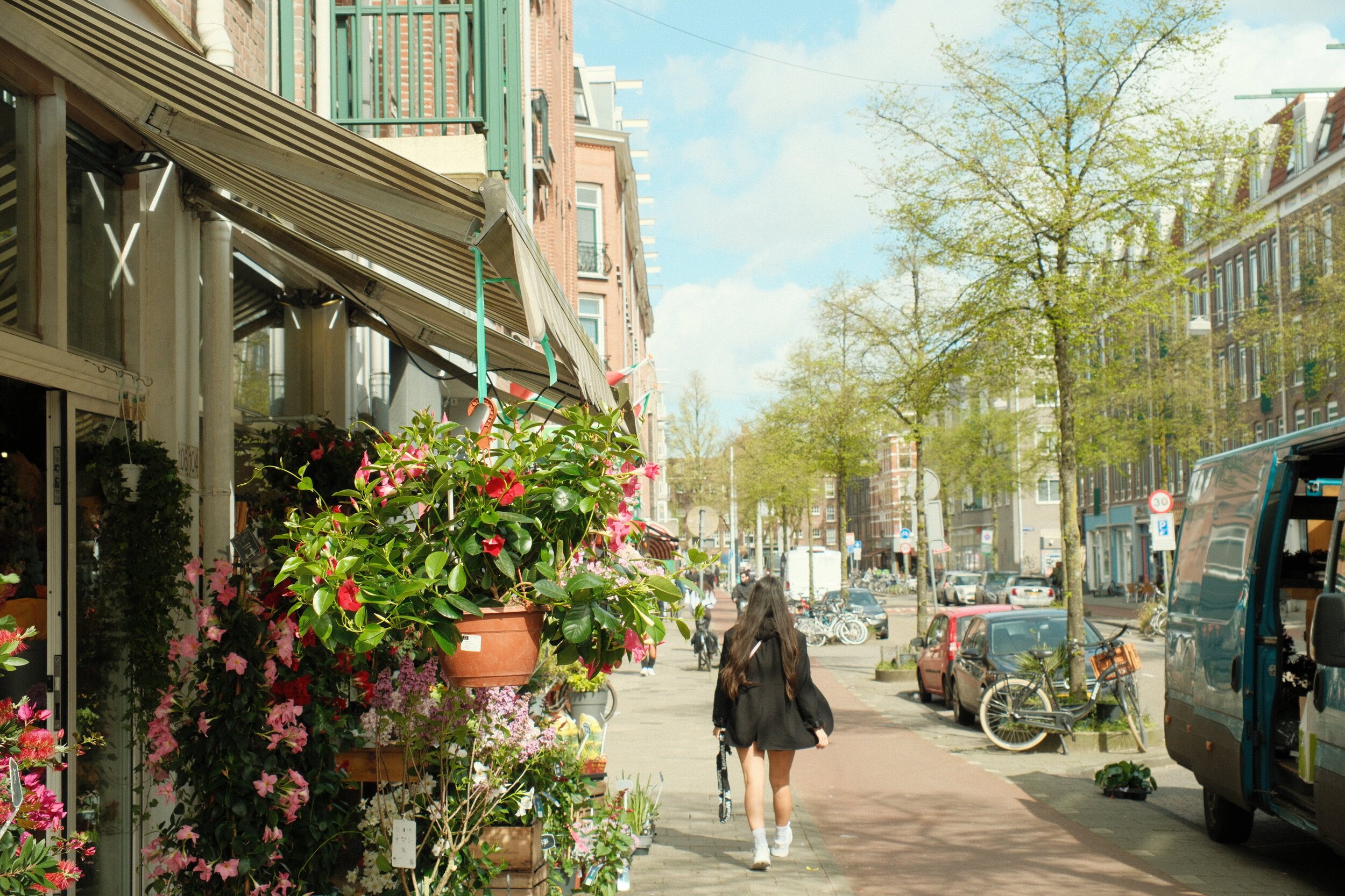 A girl walks down a city sidewalk lined with flower shops and green trees on a sunny day.