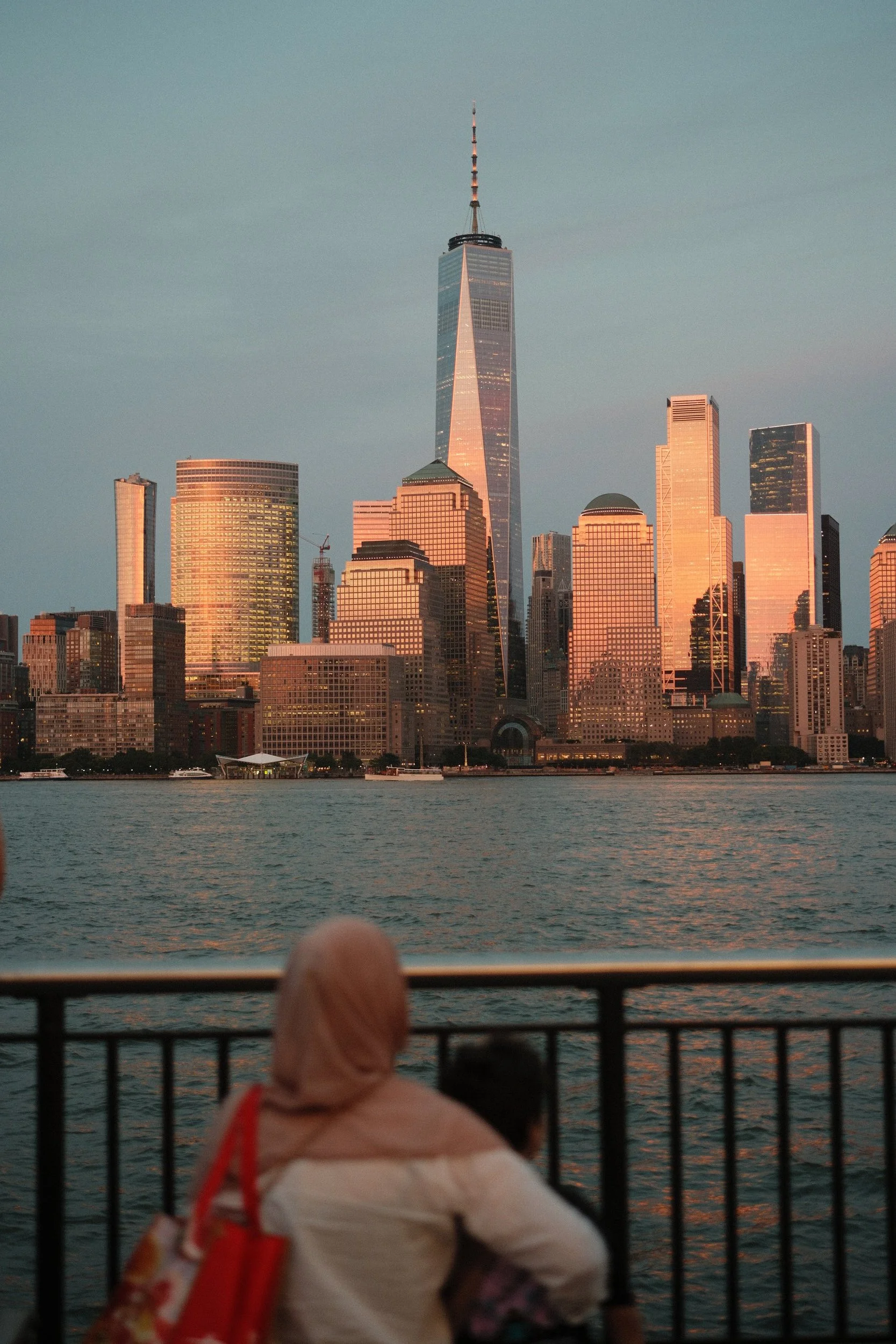 Person sitting with legs crossed on a boat or dock, overlooking the New York City skyline at sunset, including One World Trade Center