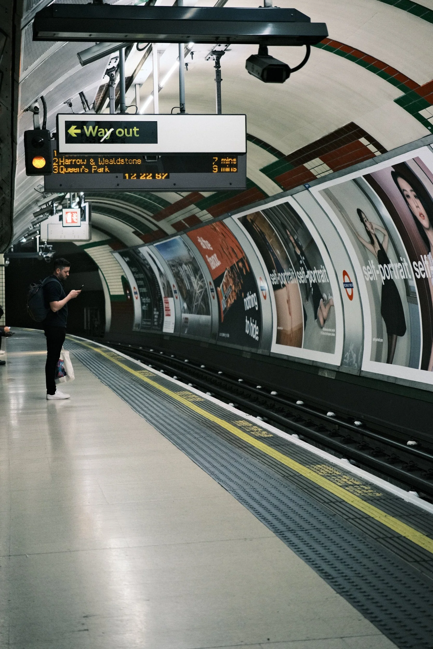 A person standing on a London Underground station platform looking at their phone, with advertising posters along the curved wall behind them and a digital sign overhead indicating train schedules.