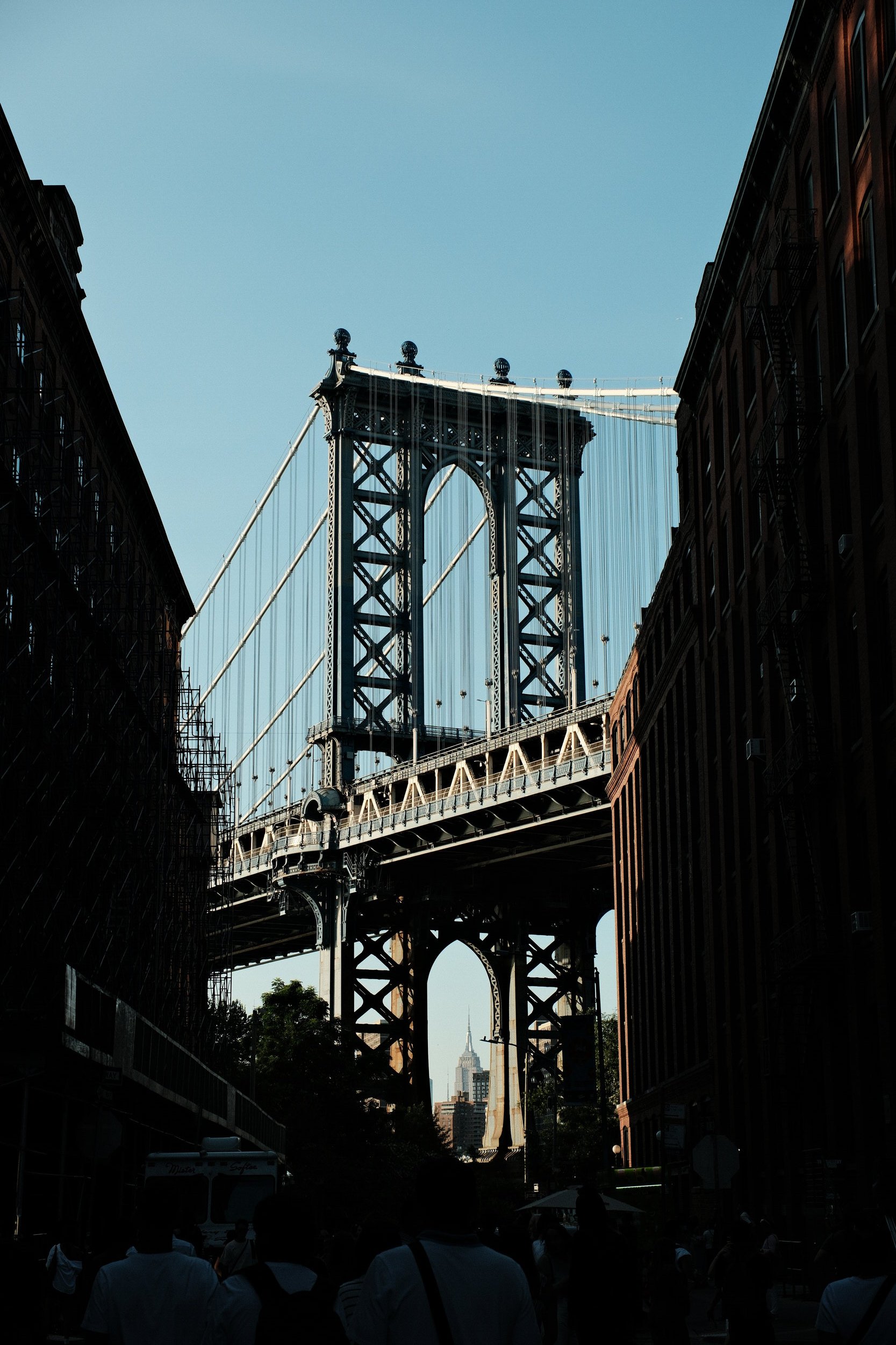 City street view with a suspension bridge framing the Empire State Building in the background, seen through a gap between buildings on a sunny day.