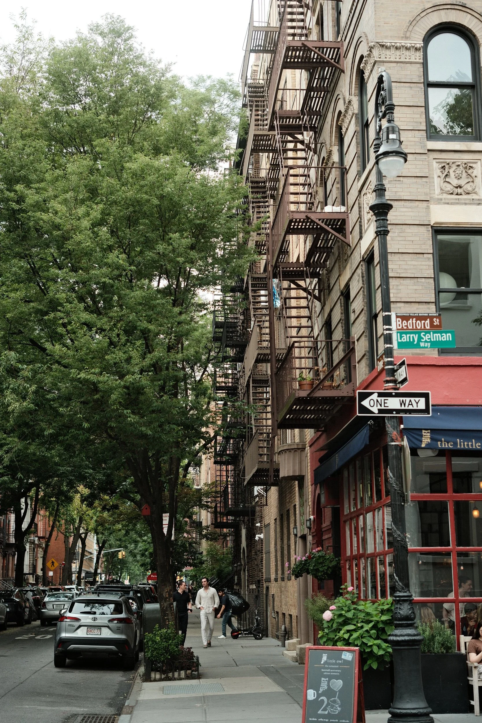 City sidewalk on Bedford Street with a brick building featuring fire escape stairs, street signs, and pedestrians walking, surrounded by trees and parked cars.