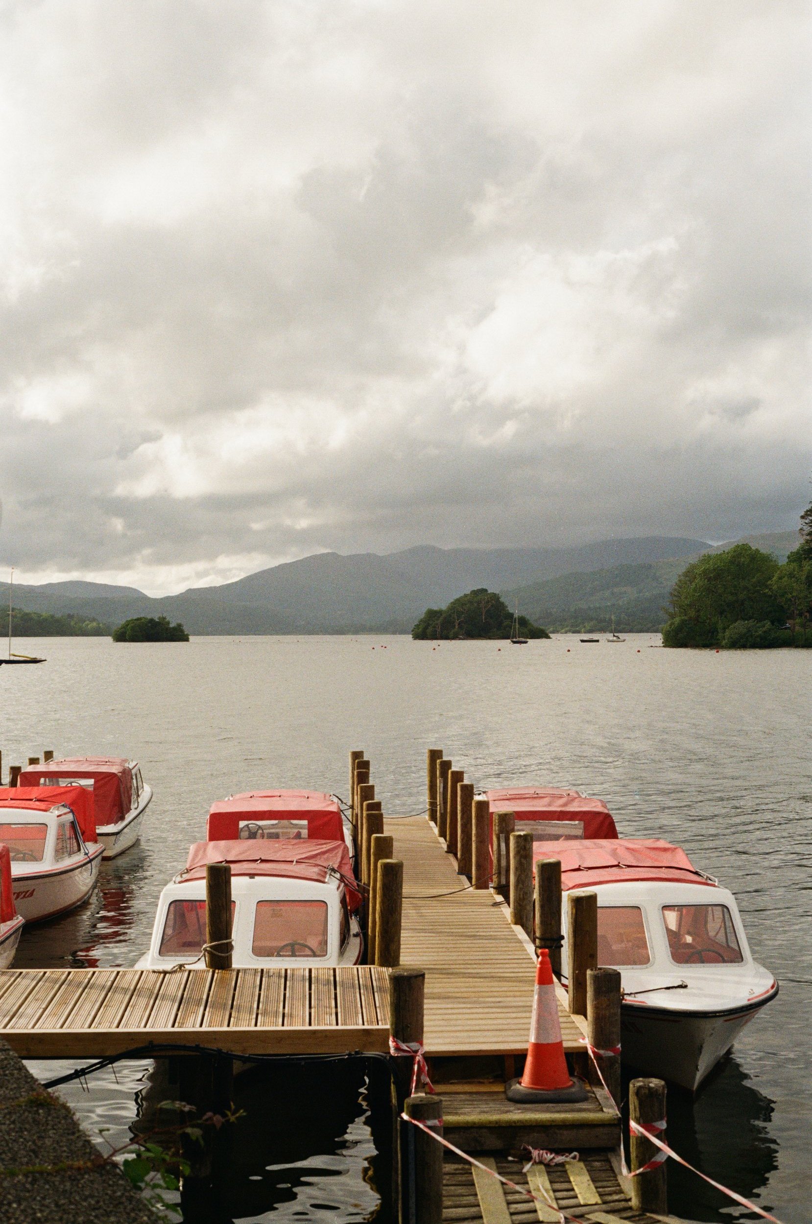 Overcast sky over a lake with a wooden dock and small boats with red covers tied to the dock, mountains in the background, and trees on the shoreline.