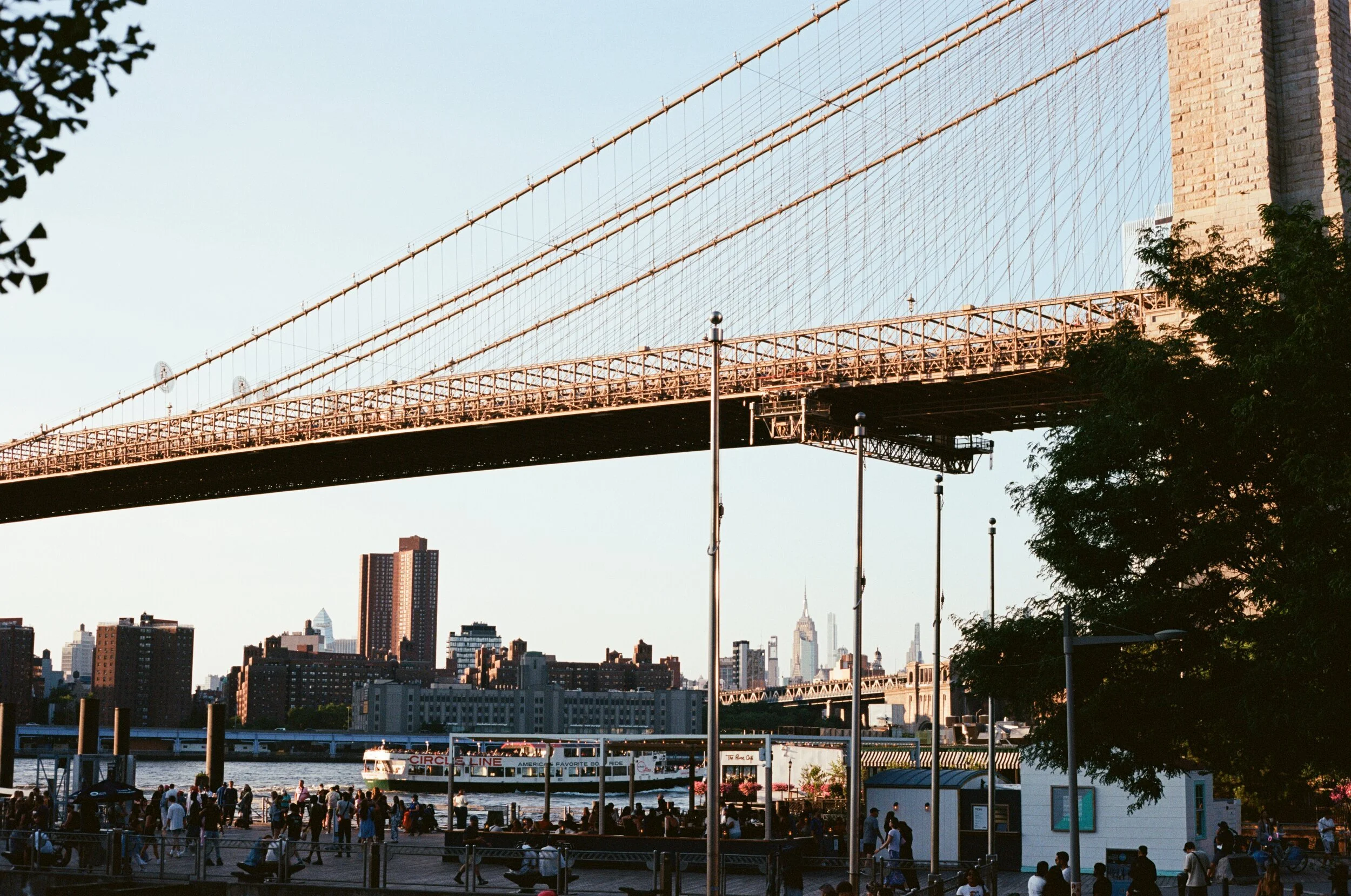 View of a bridge over New York City skyline with a river in the foreground and a crowd gathering along the shoreline.