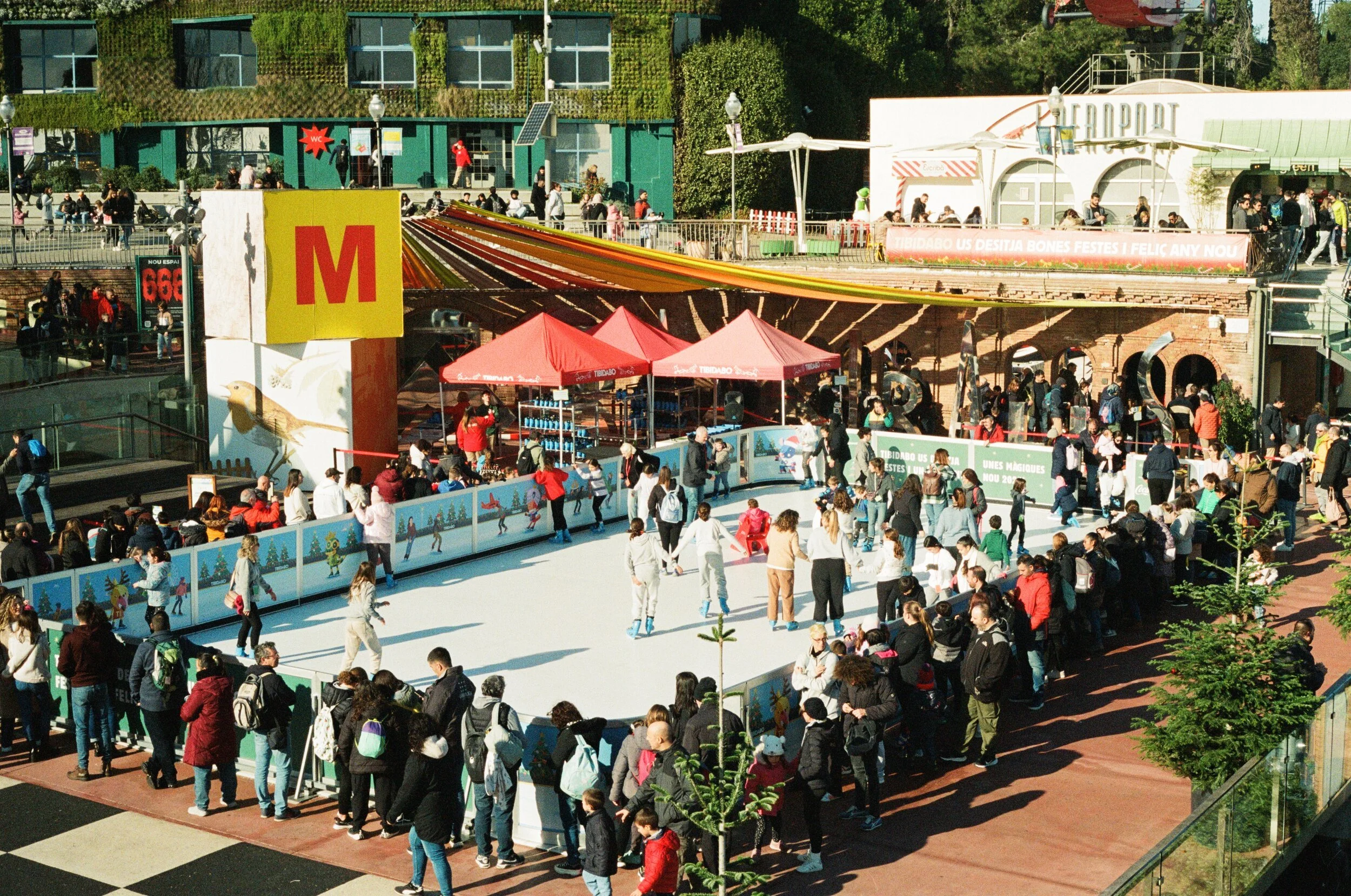Public ice skating rink with many people skating and waiting in line, surrounded by food stands, tents, and railings, with a building in the background.