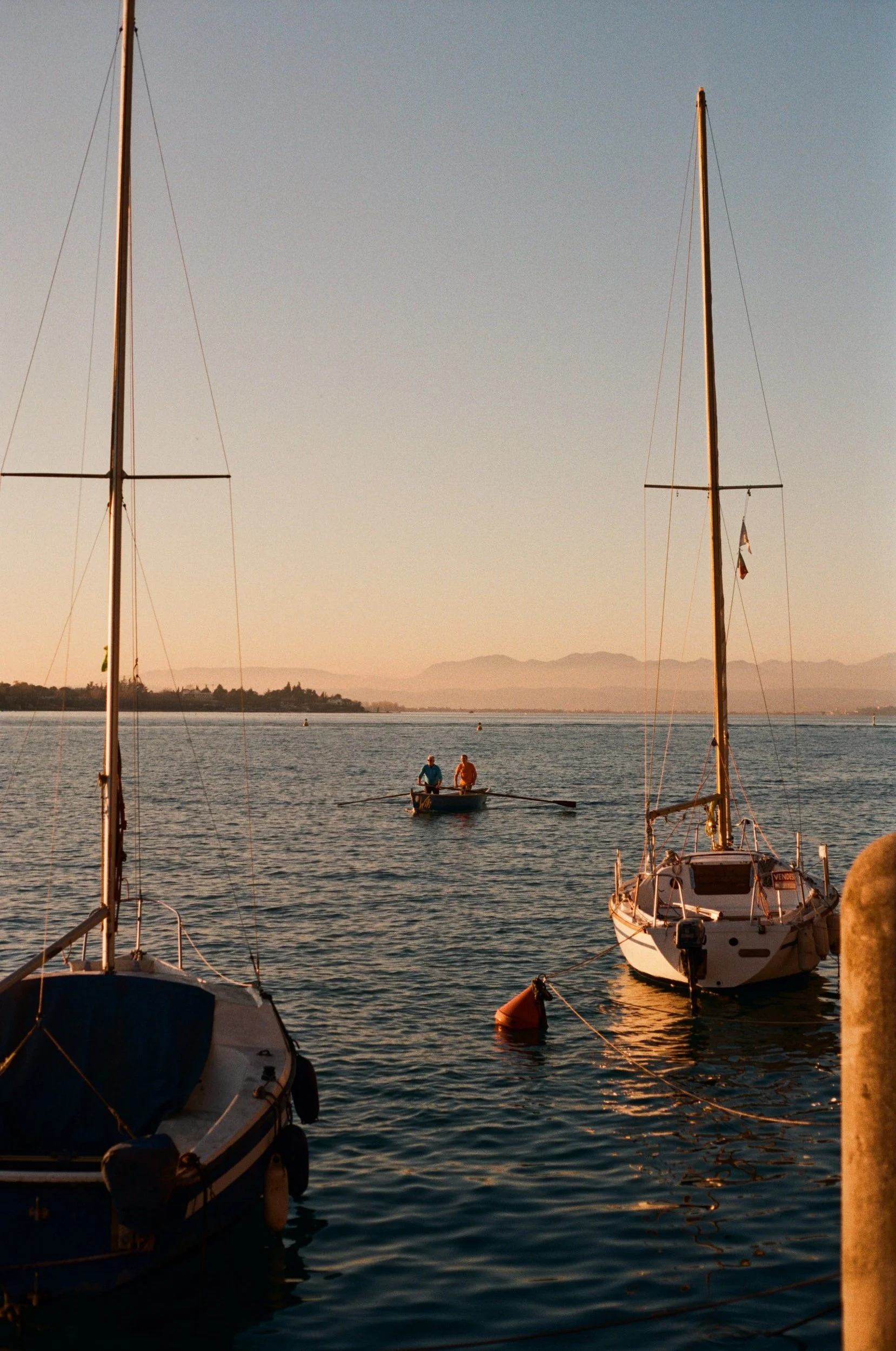 Two sailboats docked at the pier and one small boat with two people rowing in the water during sunset, with mountains in the background.