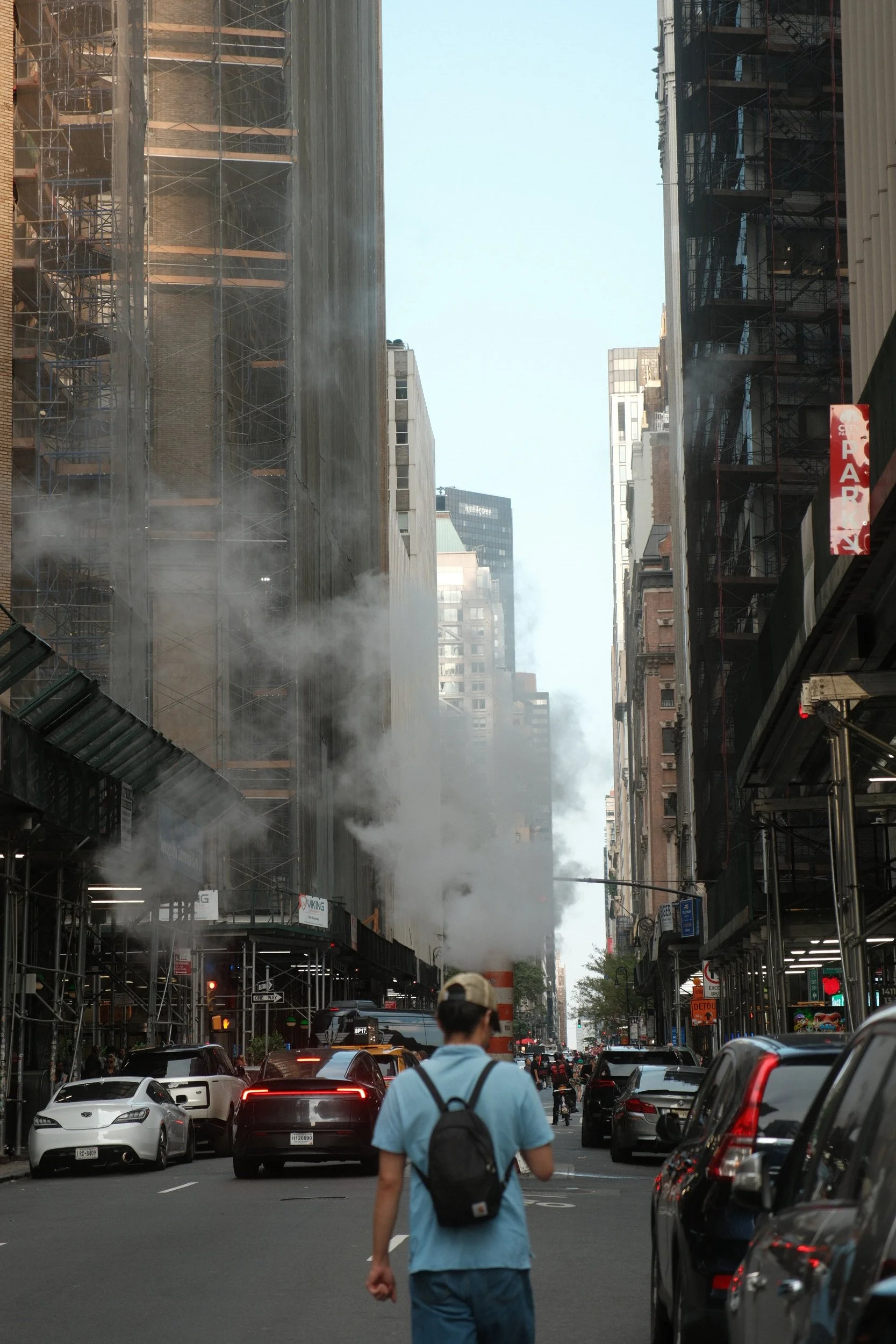 City street scene with smoke and dust rising from construction on buildings on either side, cars parked and moving, pedestrians walking, and a person with a backpack and blue shirt in the foreground.