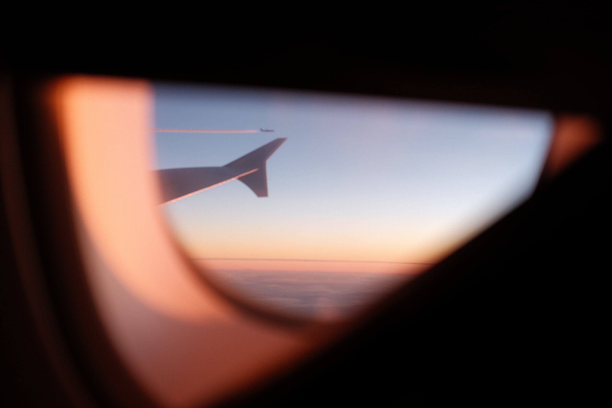 View through an airplane window showing a wing, with a sunset sky and contrail with a small airplane in the distance.