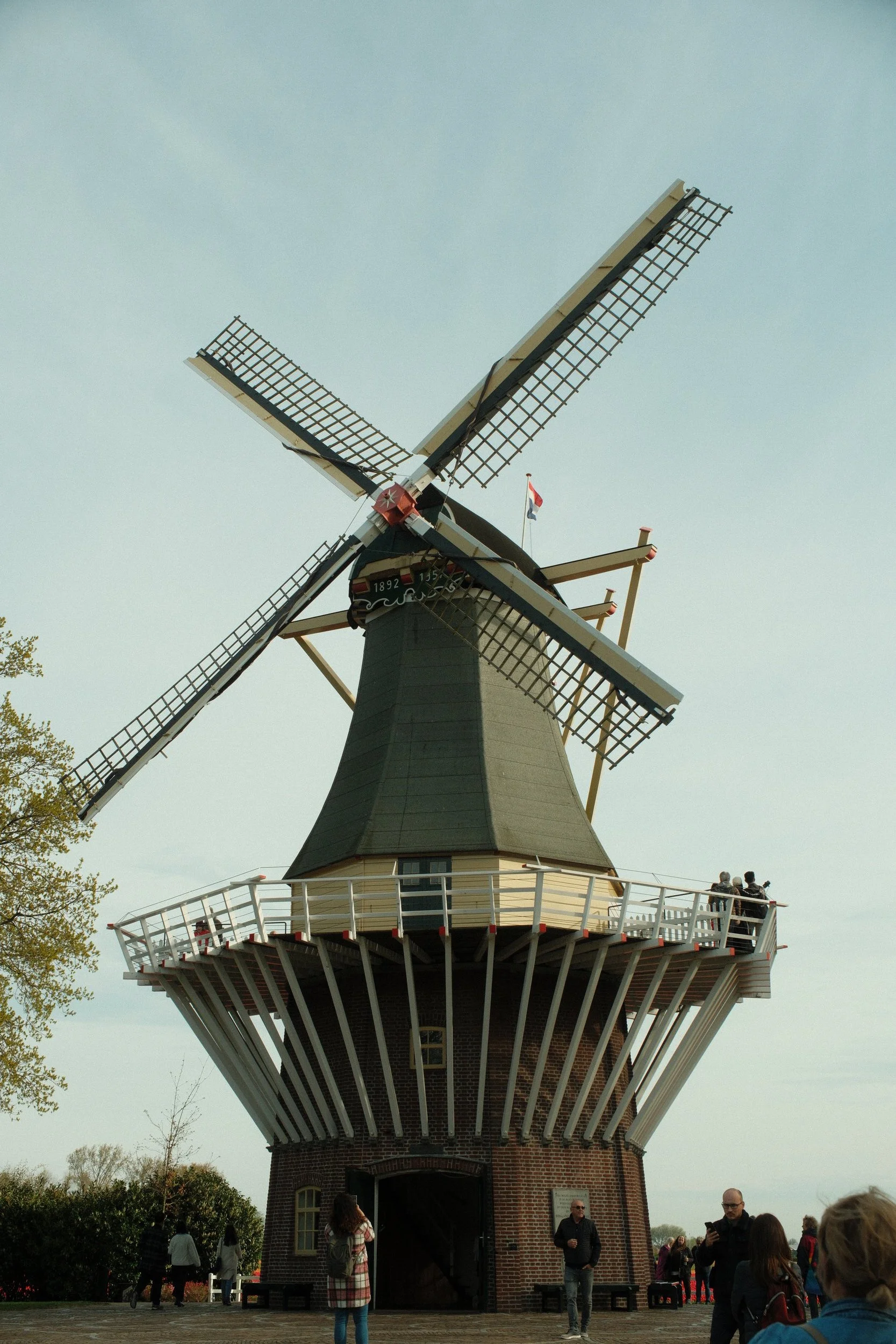 A traditional Dutch windmill with four large blades, surrounding a viewing platform with a white railing, and a brick base. People are gathered around it, some taking pictures.