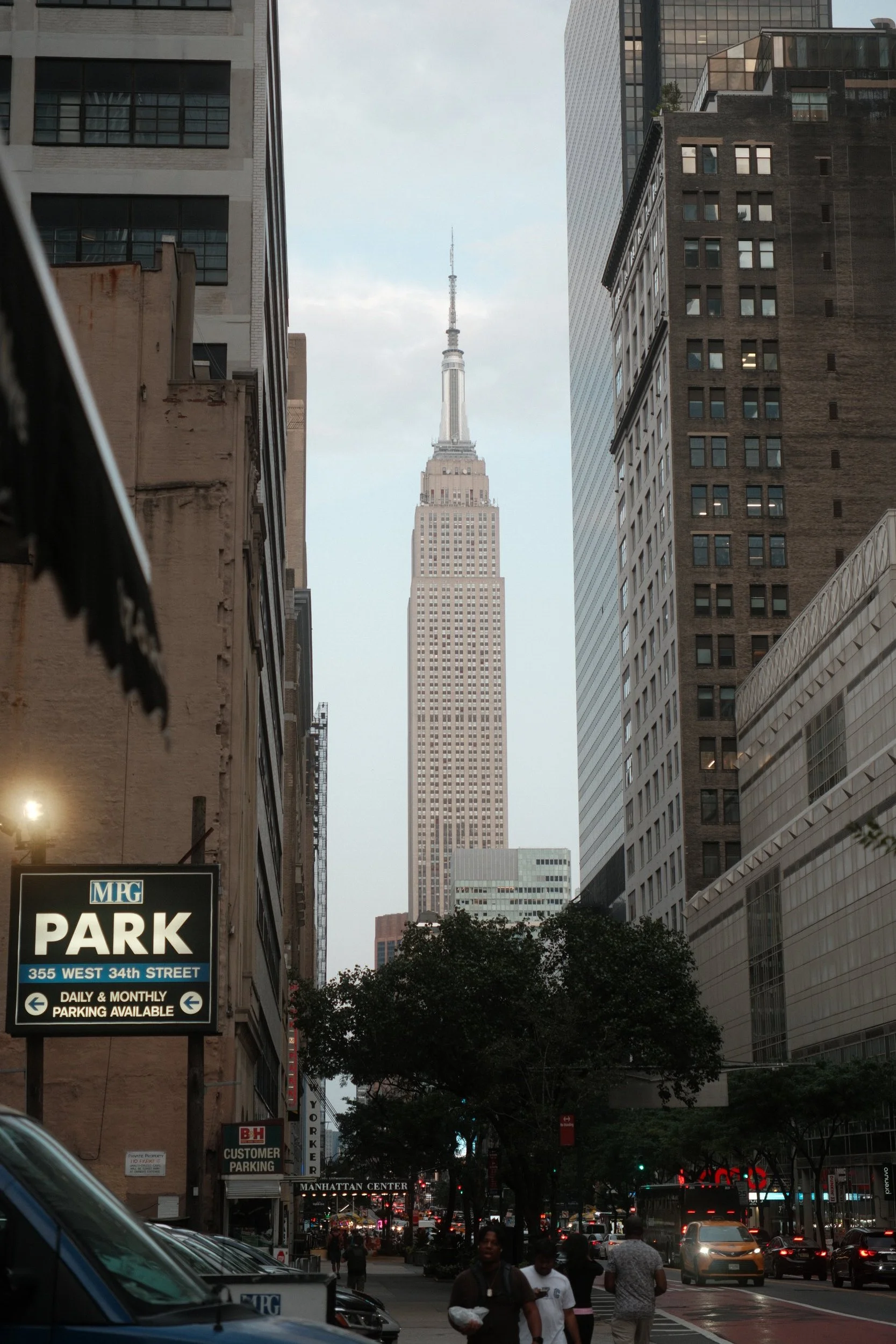 The Empire State Building is visible in the distance through a narrow gap between tall city buildings on a busy street with people walking and cars parked and moving, some storefront signs including one for a parking garage, and trees lining the side