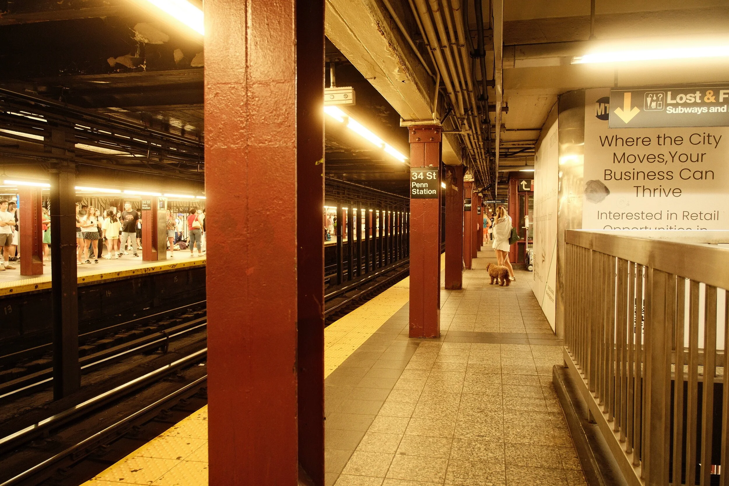 Subway station platform with people waiting for the train, red support columns, and a sign indicating 34th Street Penn Station.