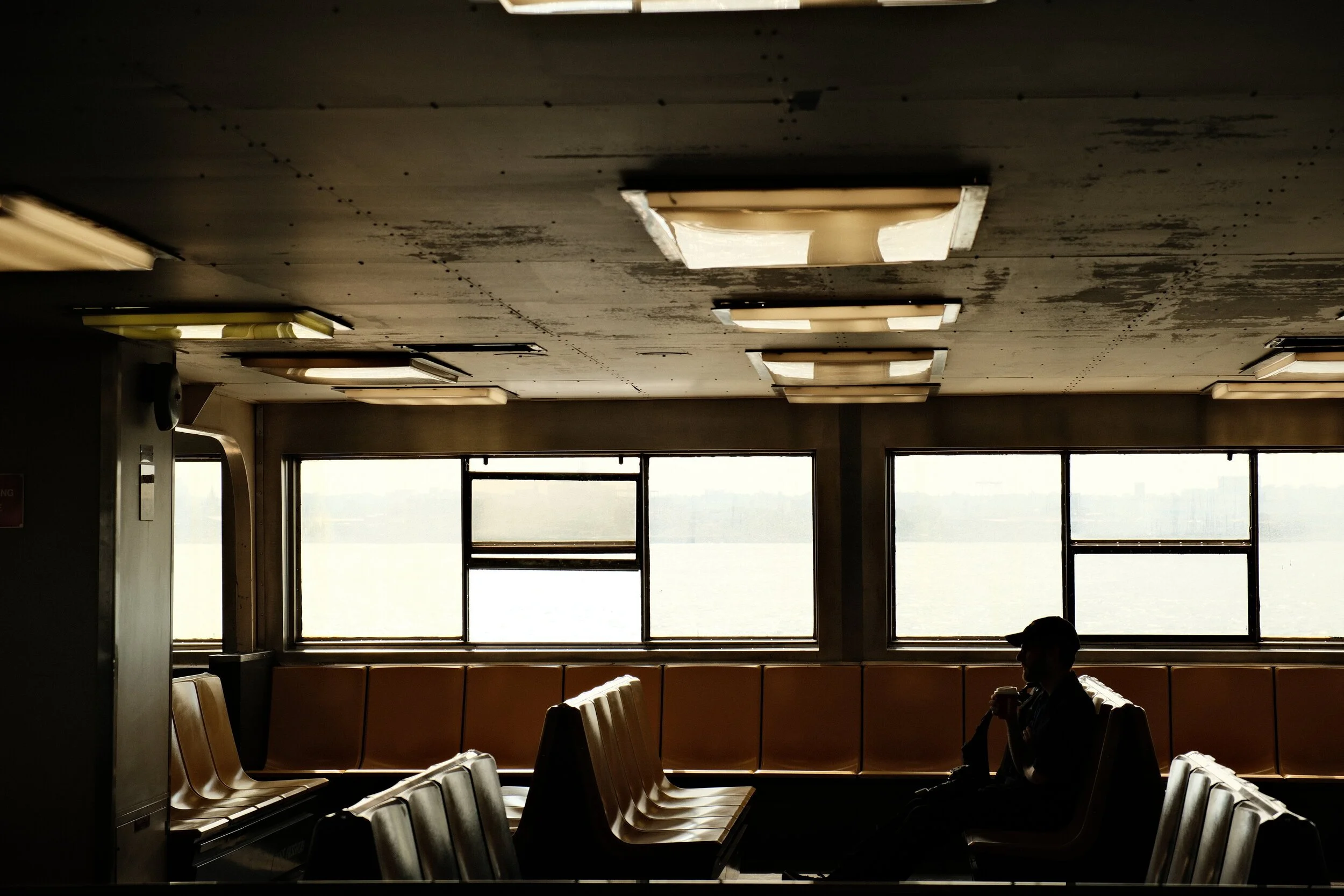 Interior view of an airport or train station waiting area with empty beige seats, large windows showing an overcast sky, and an individual sitting with a backpack.