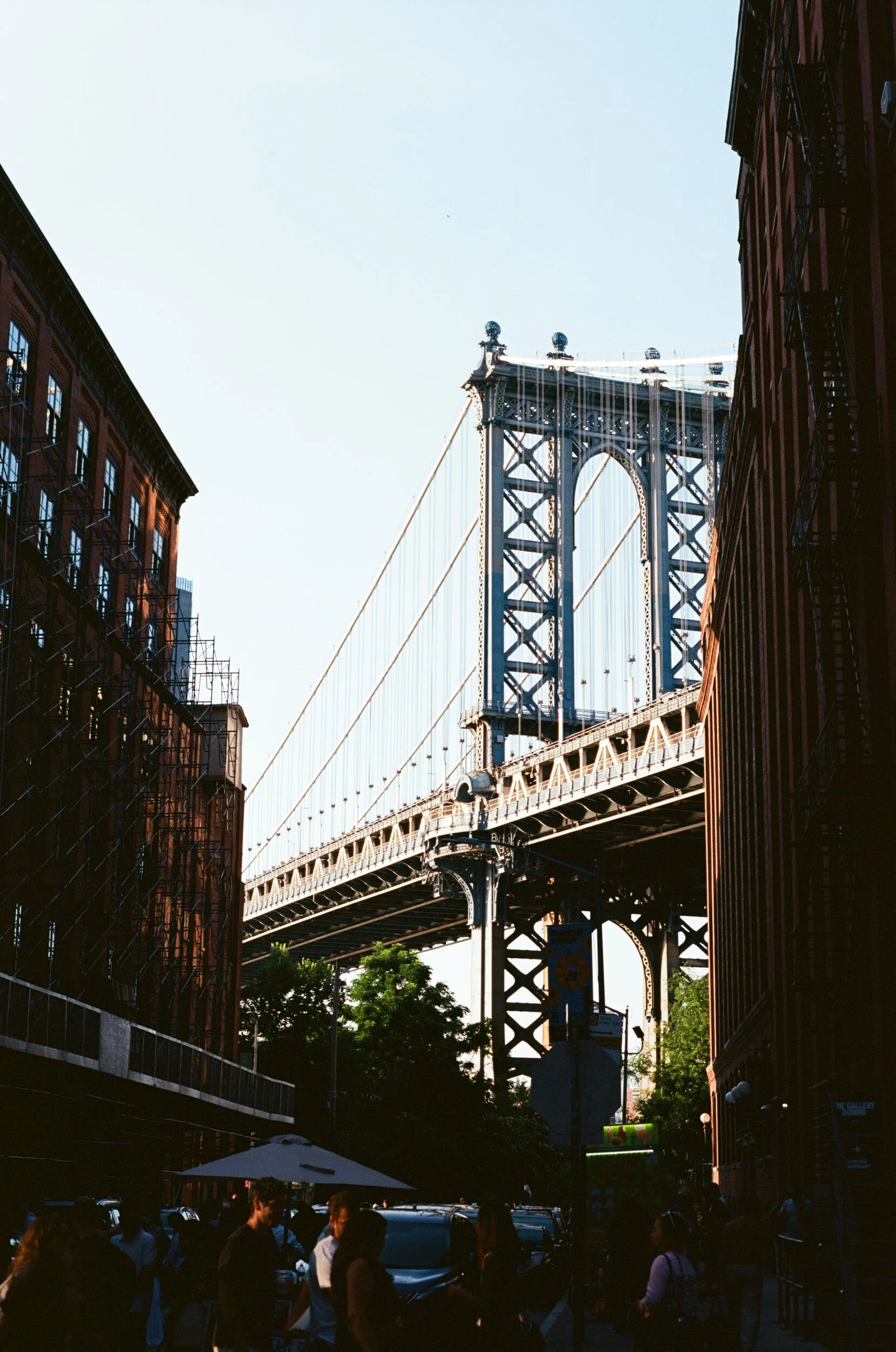 View of the Manhattan Bridge seen from a street in Brooklyn, New York City, with buildings on either side, people walking, and trees below.