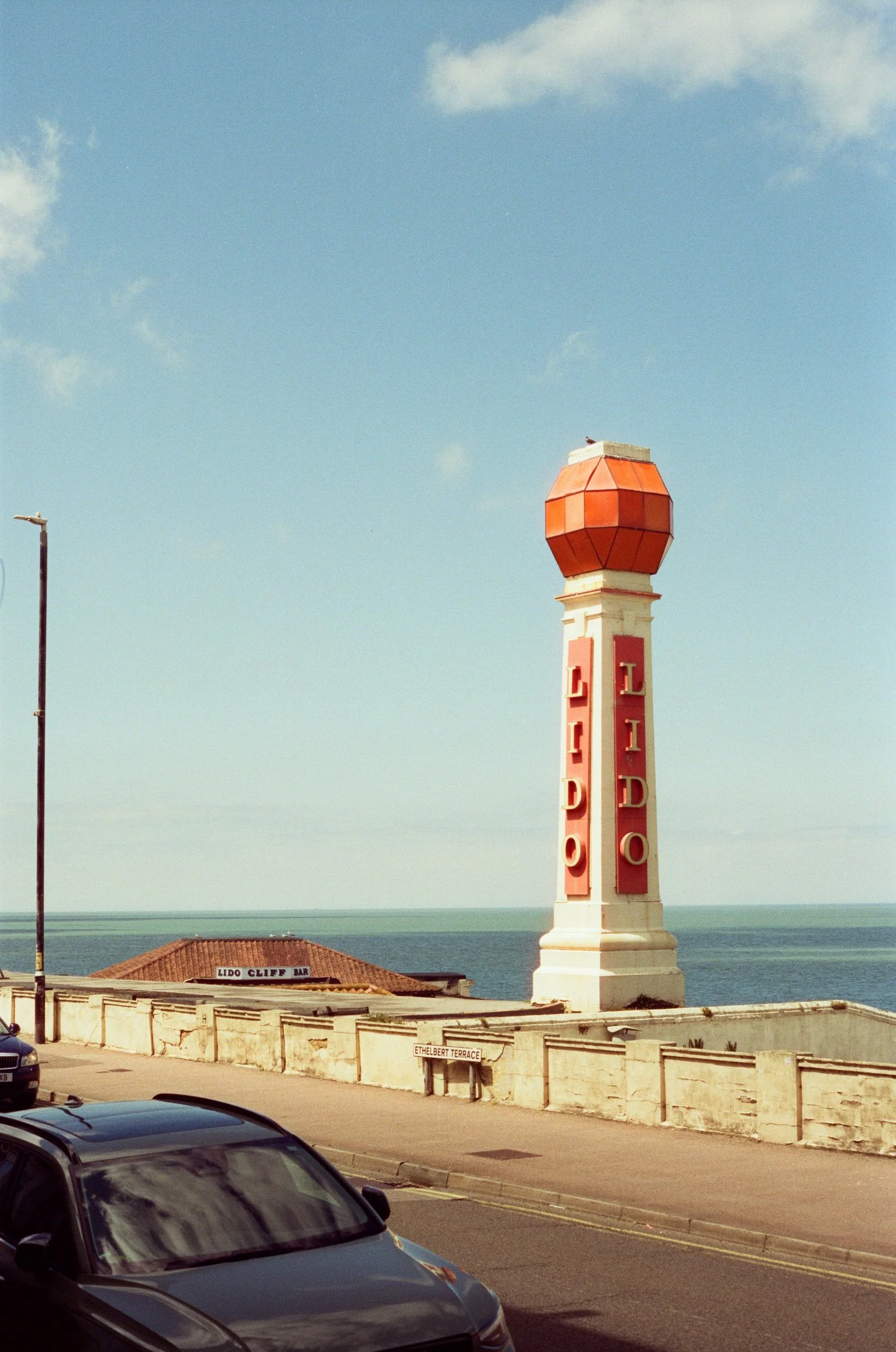 A lighthouse with the word 'LIDO' on it, situated near an ocean, with cars parked along the street and a building with a sign that says 'LIDO CLIFF BAR' in the background.