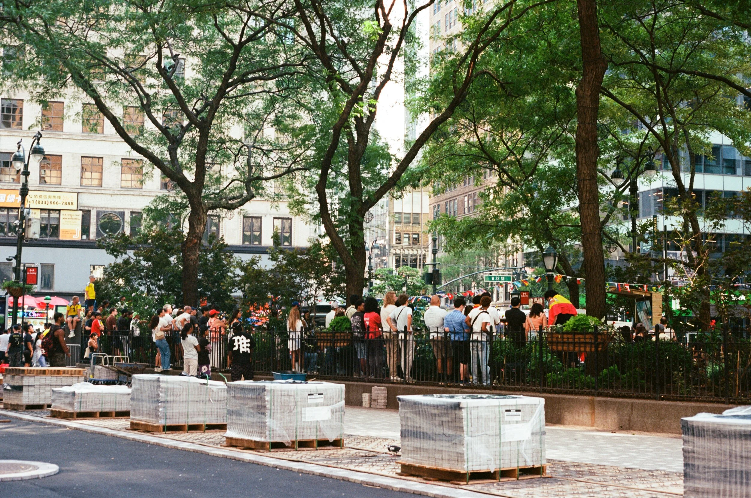 A busy city park with many people gathered, trees with green foliage, tall buildings in the background, and construction materials on the ground.