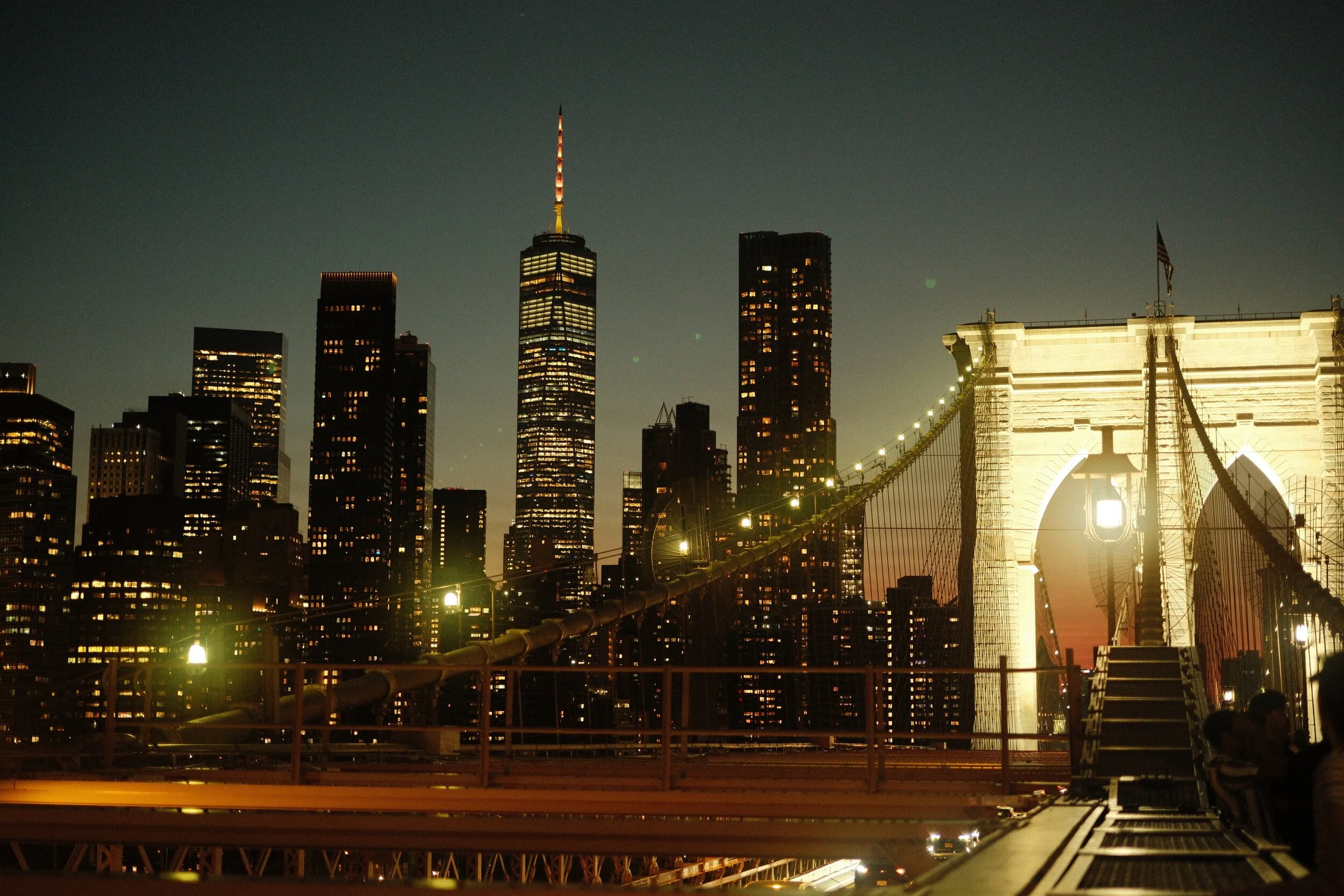 Nighttime view of the New York City skyline featuring the iconic Brooklyn Bridge illuminated with lights and the One World Trade Center building lit up in the background.