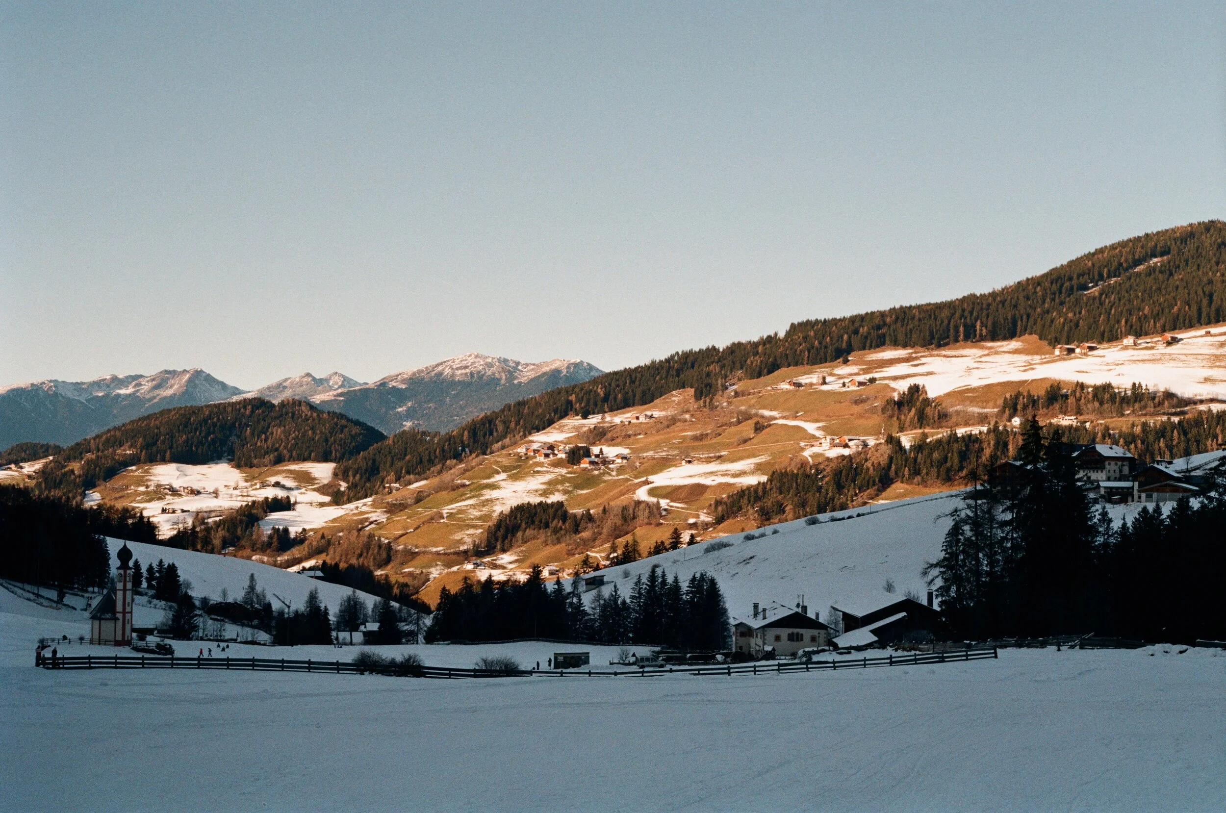 Scenic view of snow-covered fields and houses in a mountainous landscape during winter, with rolling hills and distant snow-capped peaks under a clear blue sky.