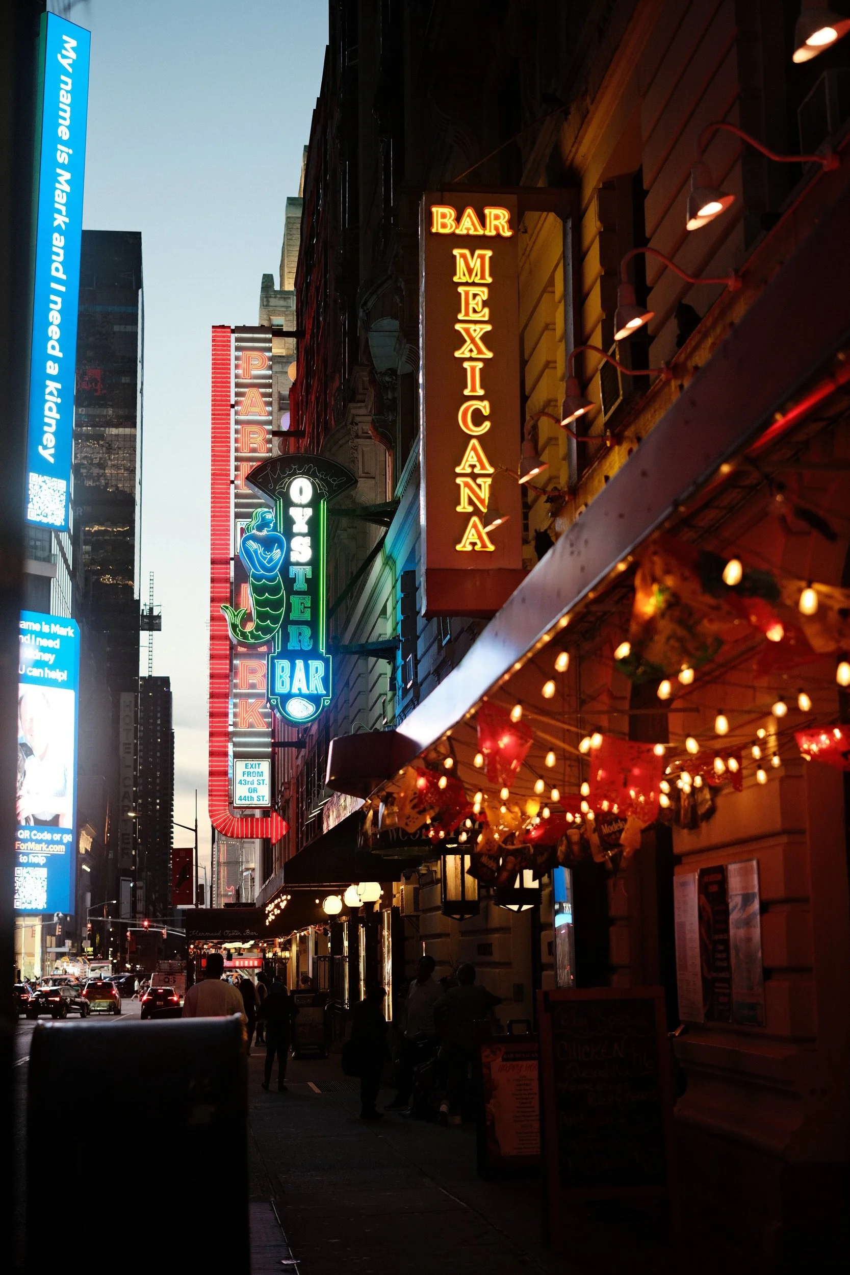 Nighttime view of a city street with illuminated signs for bars and restaurants, including a neon sign for 'Bar Mexican,' a sign for 'Oyster Bar,' and a digital billboard on the left side of the image, with people walking and cars on the street.