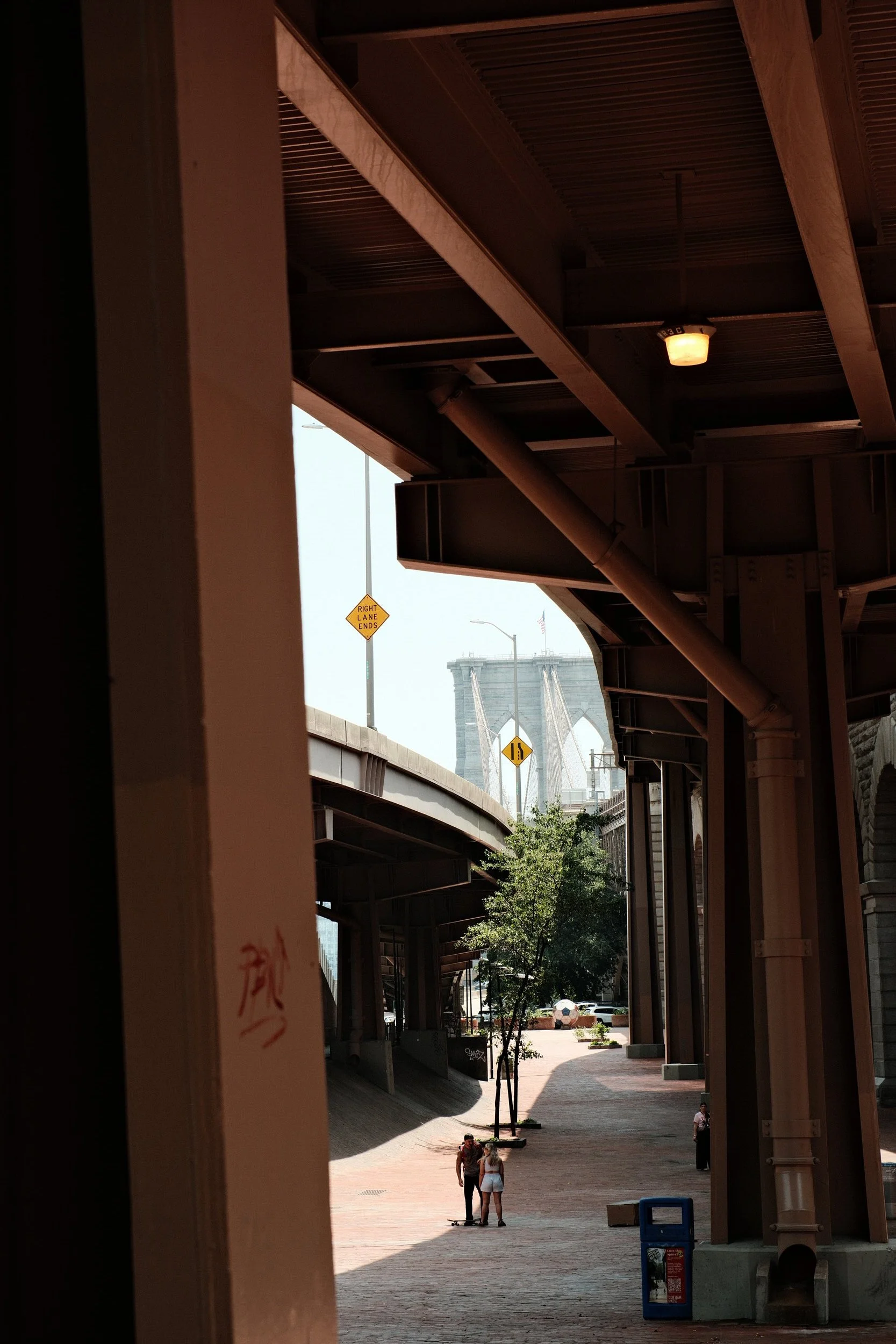 People walking and talking under an overpass with the Brooklyn Bridge visible in the background in New York City.