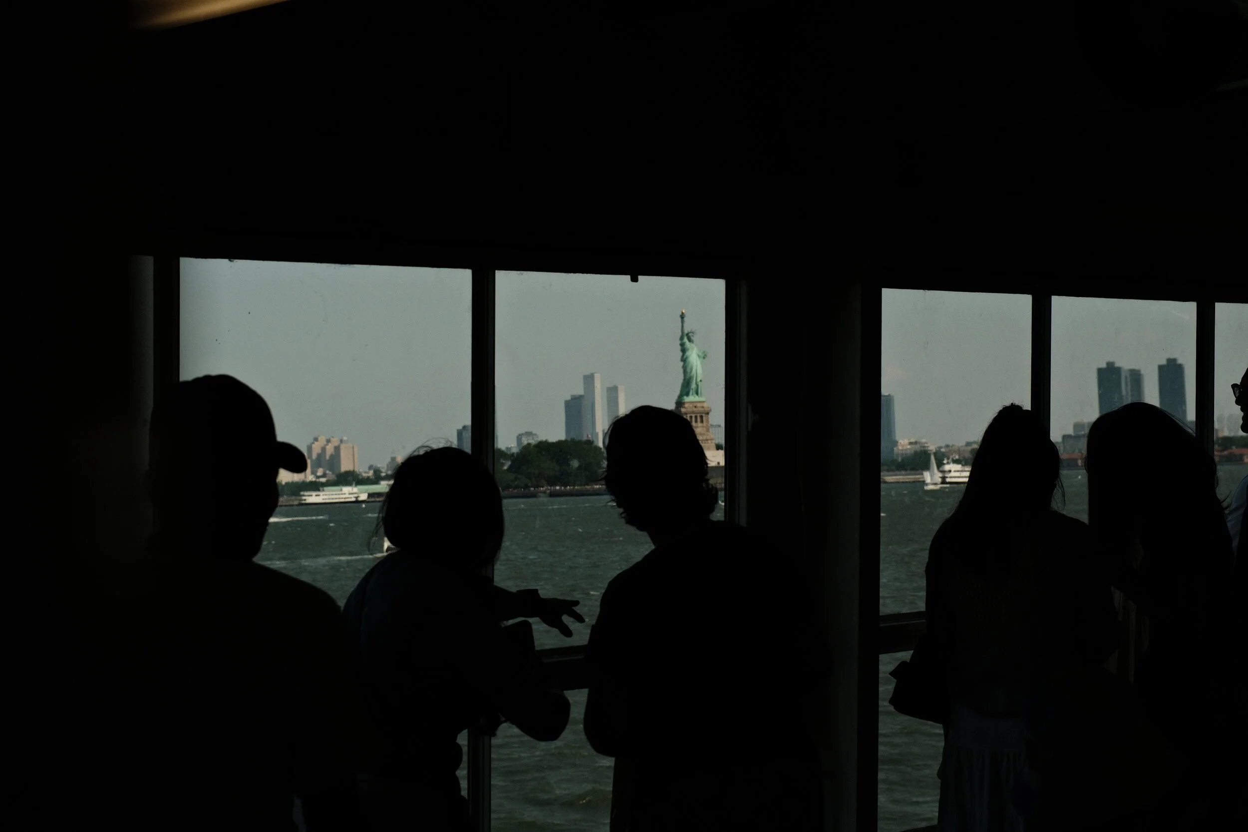 View of the Statue of Liberty seen through a window on a boat with silhouettes of people inside, and a city skyline in the background.