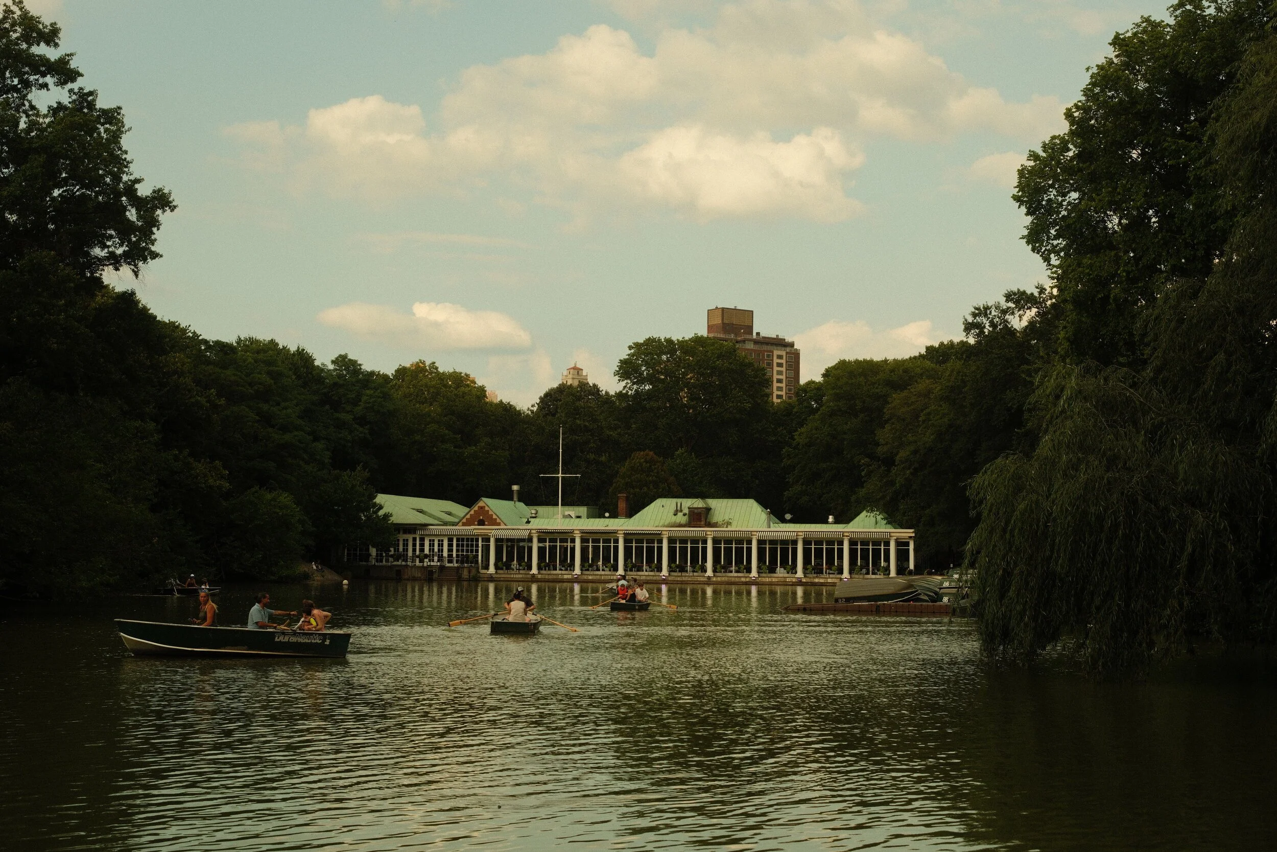 People are boating on a lake surrounded by trees, with a white building with a green roof and tall windows on the shore. Tall buildings are visible in the background under a partly cloudy sky.