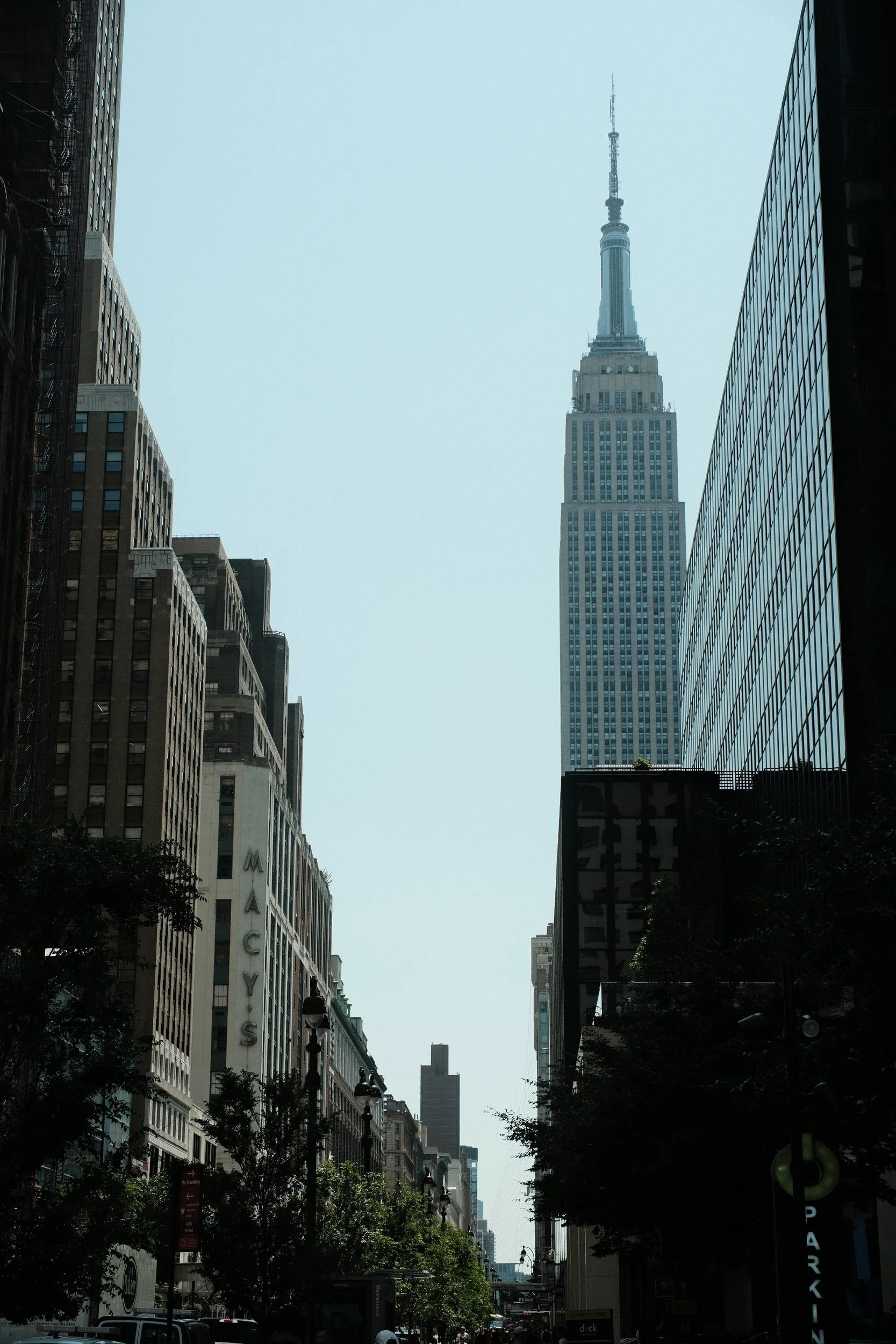 Street view in New York City with the Empire State Building visible in the distance, flanked by tall office buildings on either side, and a bustling street scene below.
