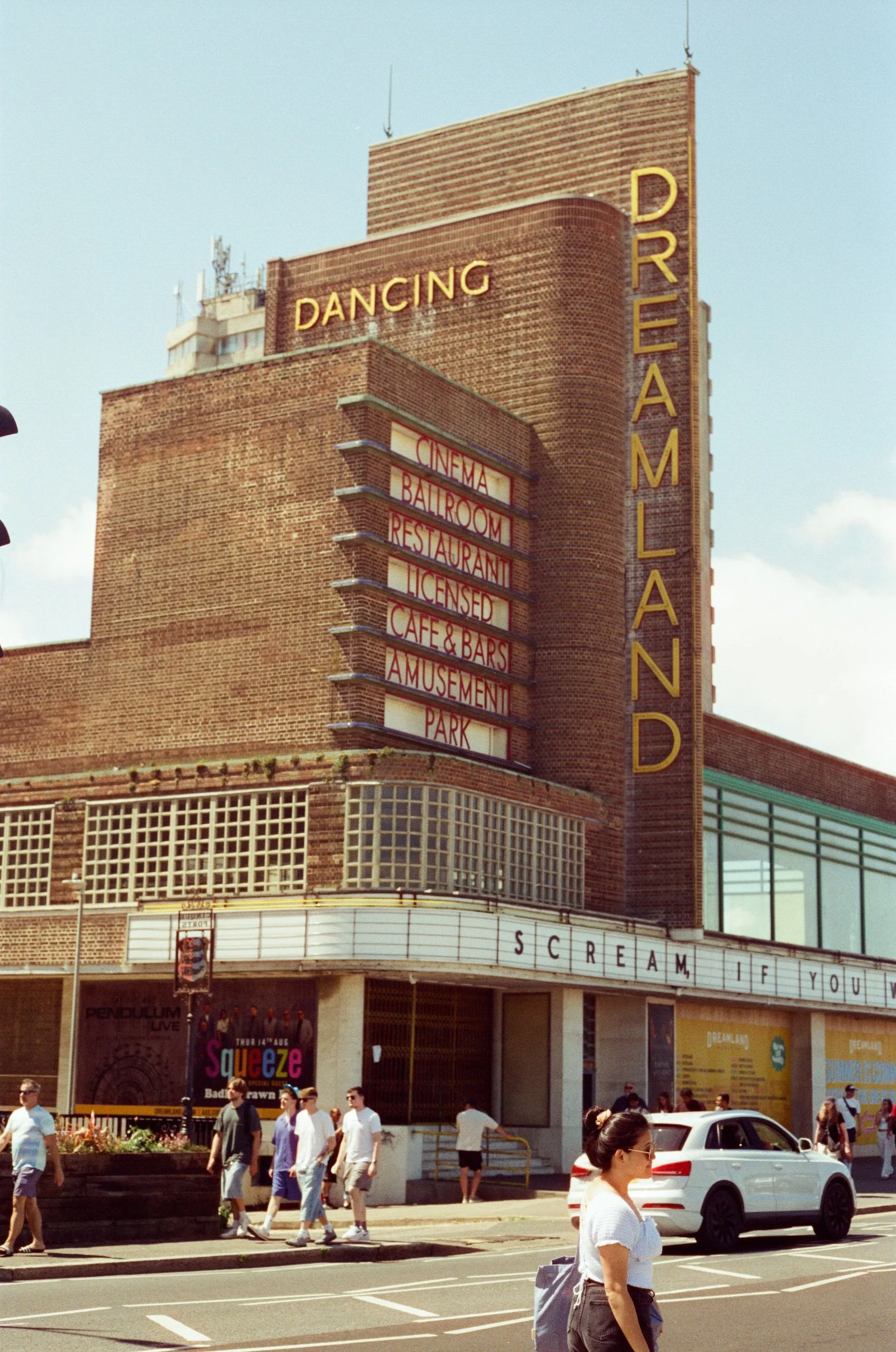 The Dreamland building with neon signs indicating it offers dancing, cinema, ballroom, restaurant, licensed venue, cafe, bars, amusement park, and a marquee displaying a show called 'Scream if You'. Pedestrians walk by and a white car drives on the s