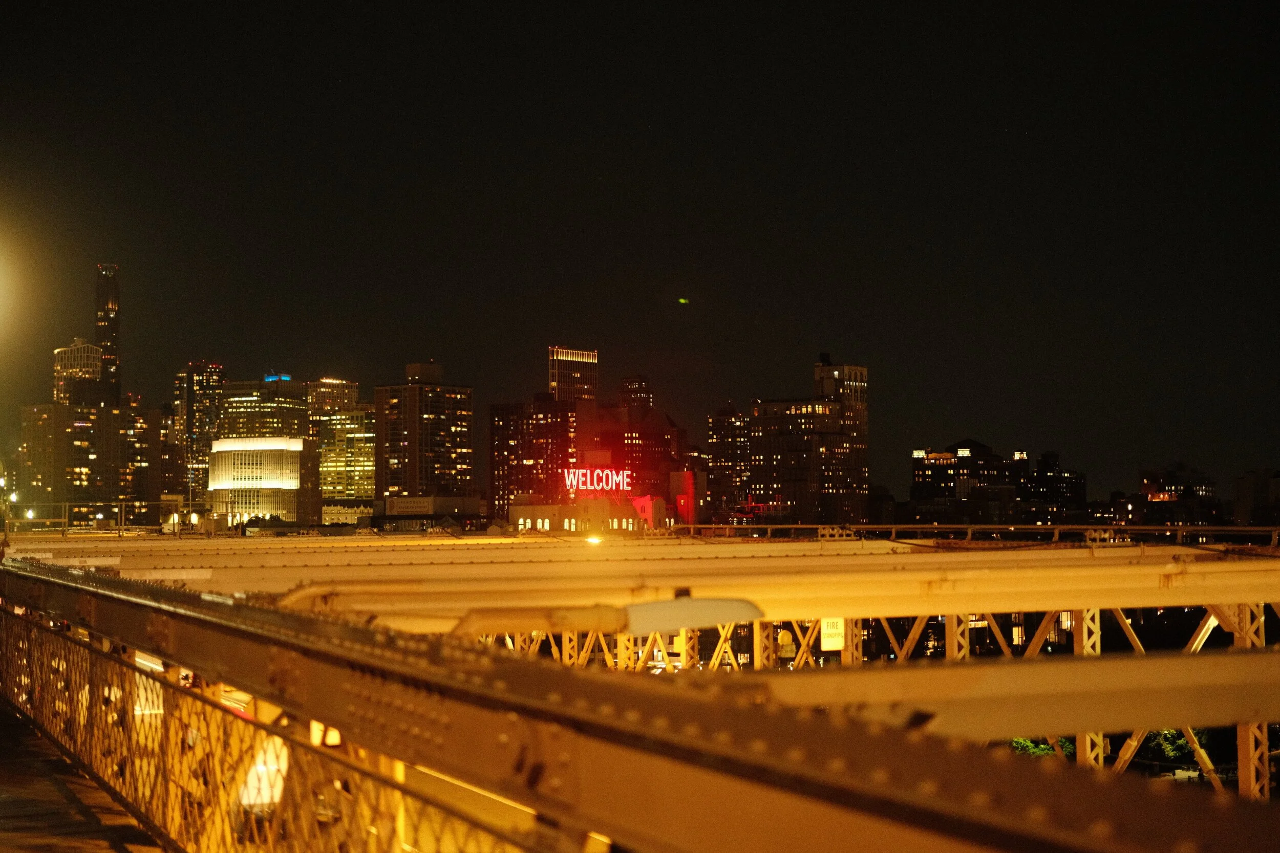 Nighttime city skyline with illuminated buildings, a prominent red "WELCOME" sign, and a view from a bridge with a metal railing in the foreground.