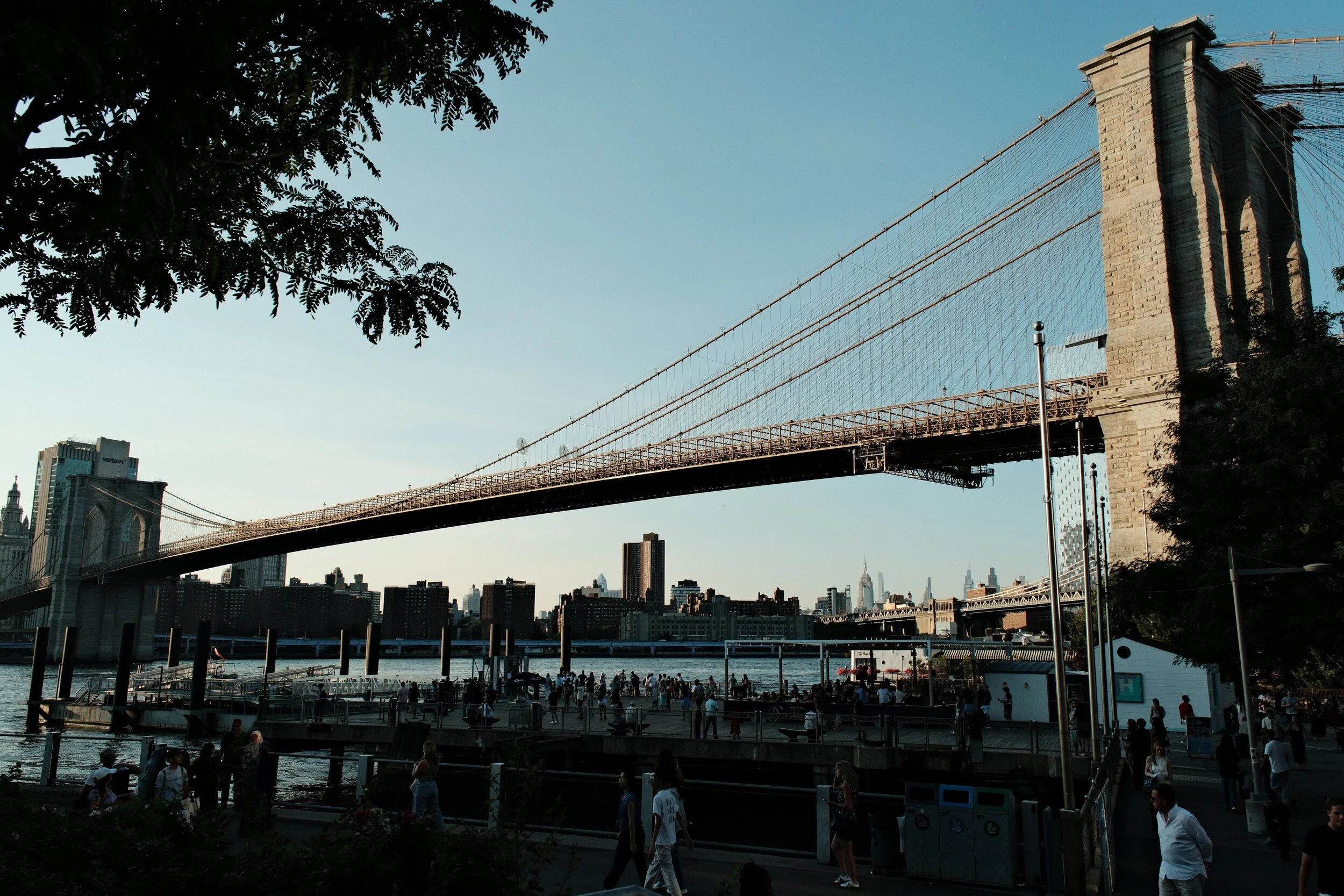 View of the Brooklyn Bridge with pedestrians walking along the walkway, City Hall Park and the New York City skyline in the background, and trees framing the scene.