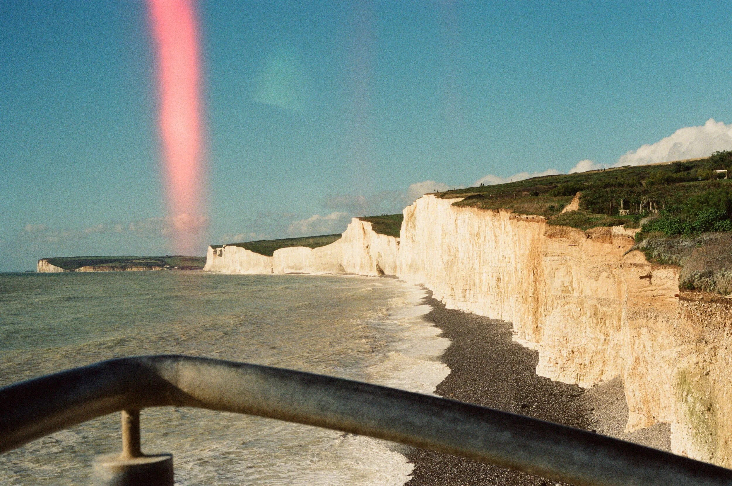 White chalk cliffs along a coastline with the sea below and a blue sky with clouds above, view from a boat deck with a railing in the foreground.