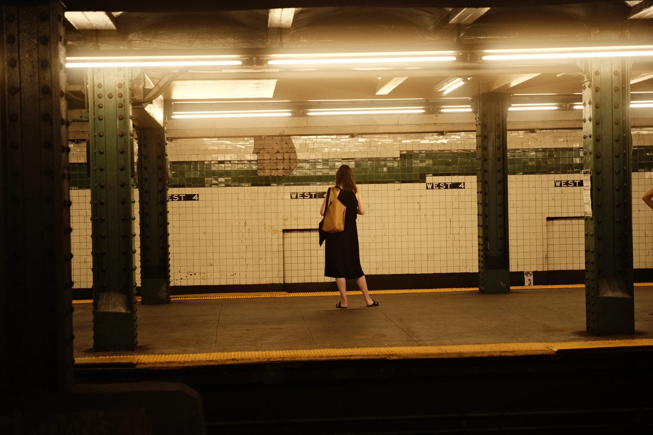 A woman with a brown backpack, black dress, and sandals standing on a subway platform, waiting near the 'West 4' subway station sign. The platform has a tiled wall and metal support beams.