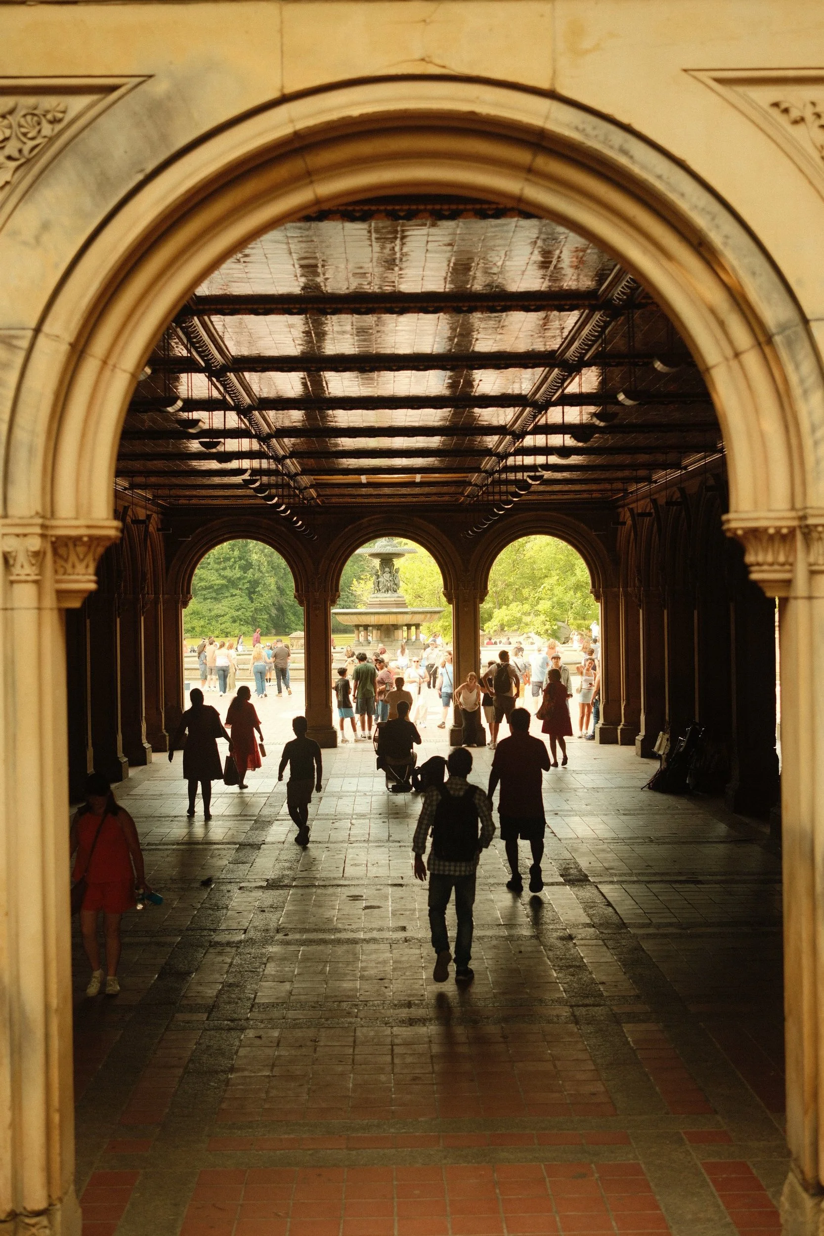 People walking through a large arched open-air corridor leading to a park with trees and a fountain in the background.