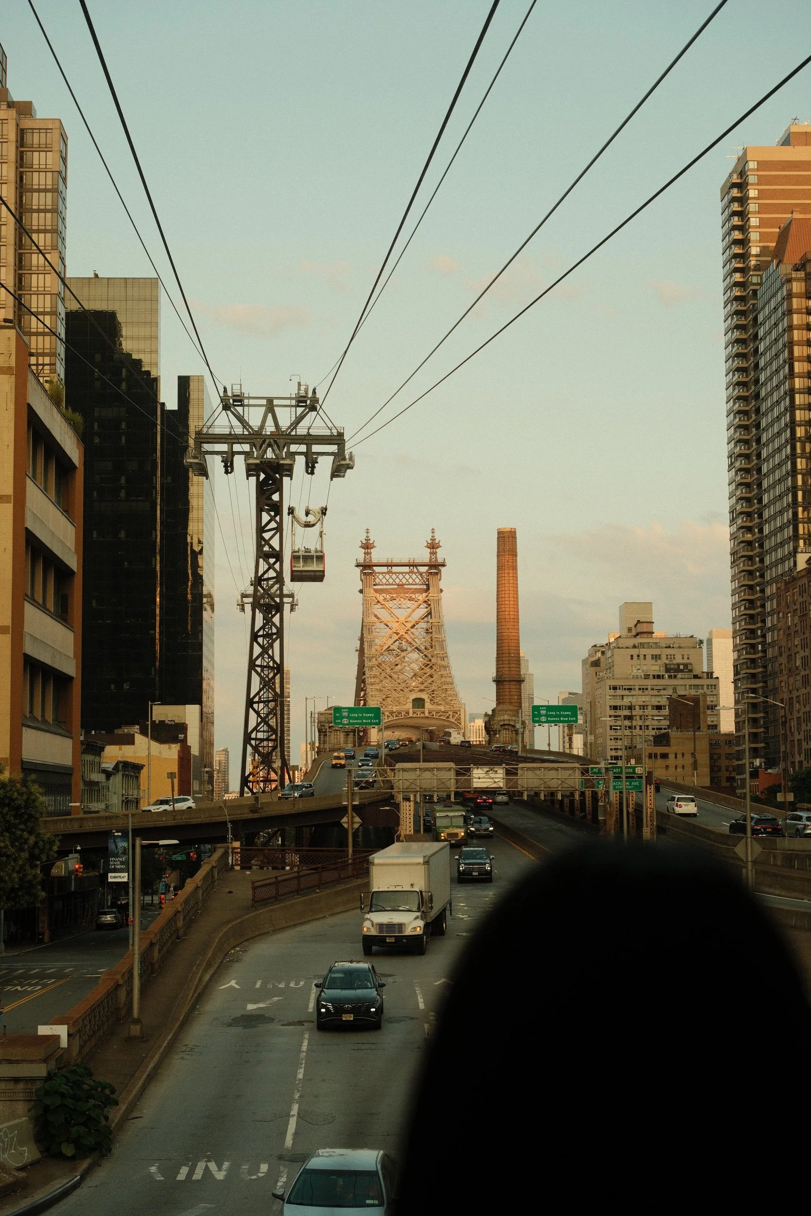 City street scene with cars driving on an elevated highway, tall buildings on either side, power lines overhead, and a bridge in the background.