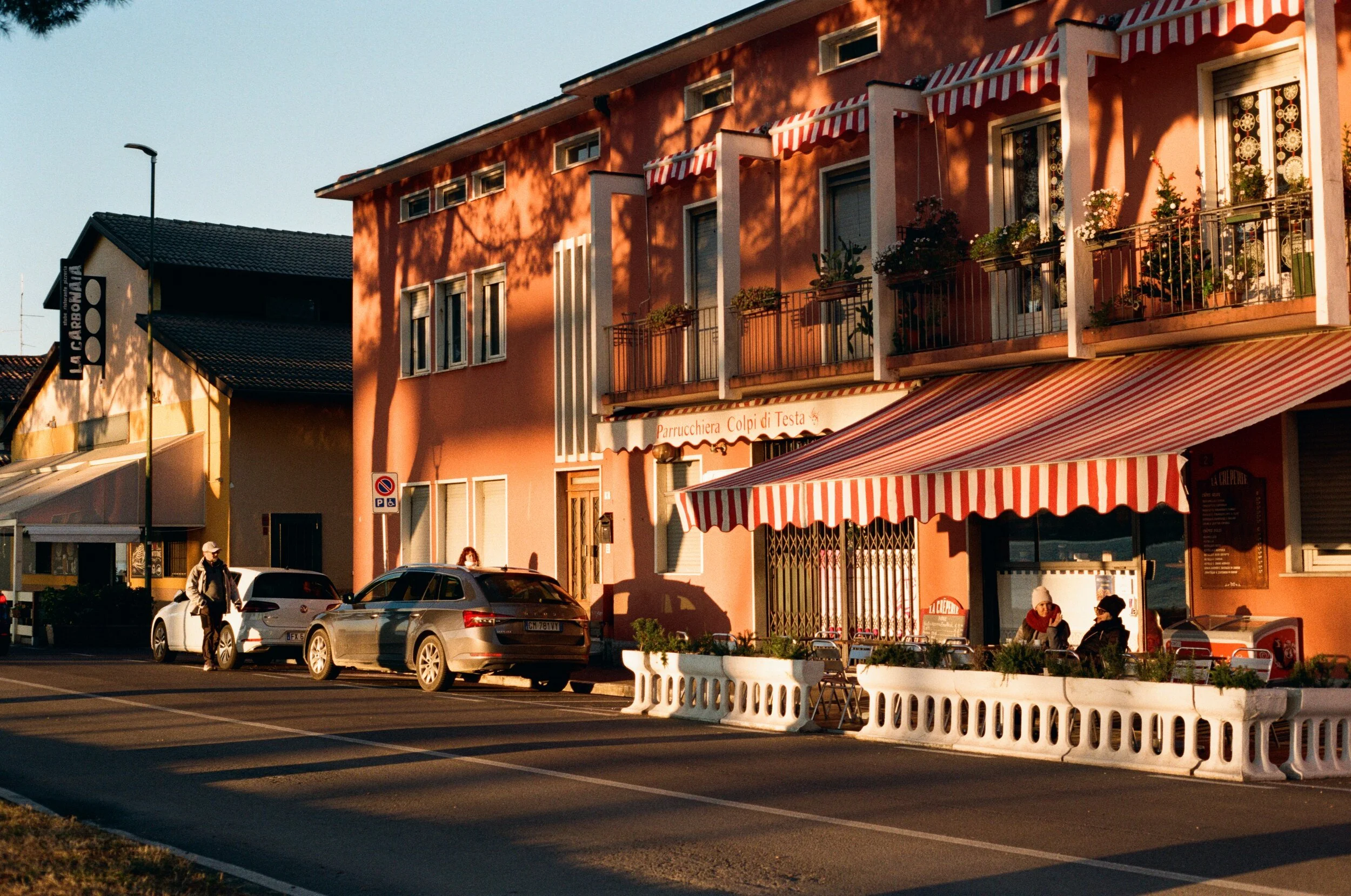 A cozy street scene during sunset with a pink building featuring striped awnings and balconies with flowers. Two people sit outside at a cafe under a red and white striped awning, and a person walks past parked cars on the street.