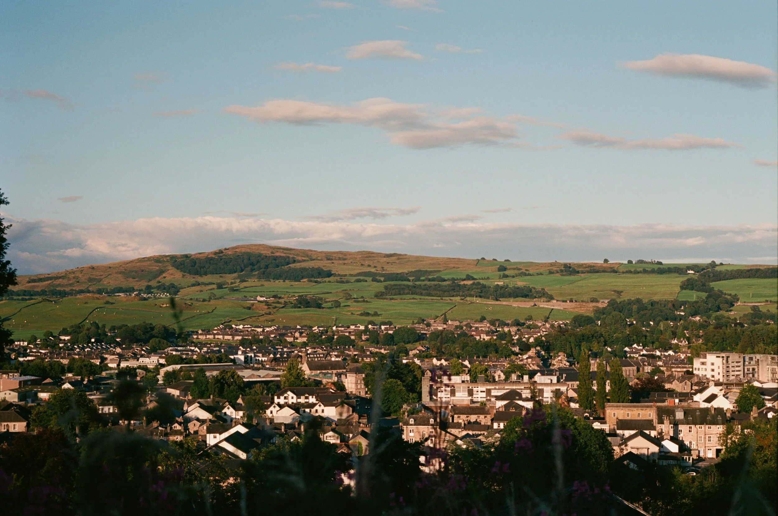 A scenic view of a small town with houses and buildings, surrounded by green hills and a blue sky with scattered clouds.