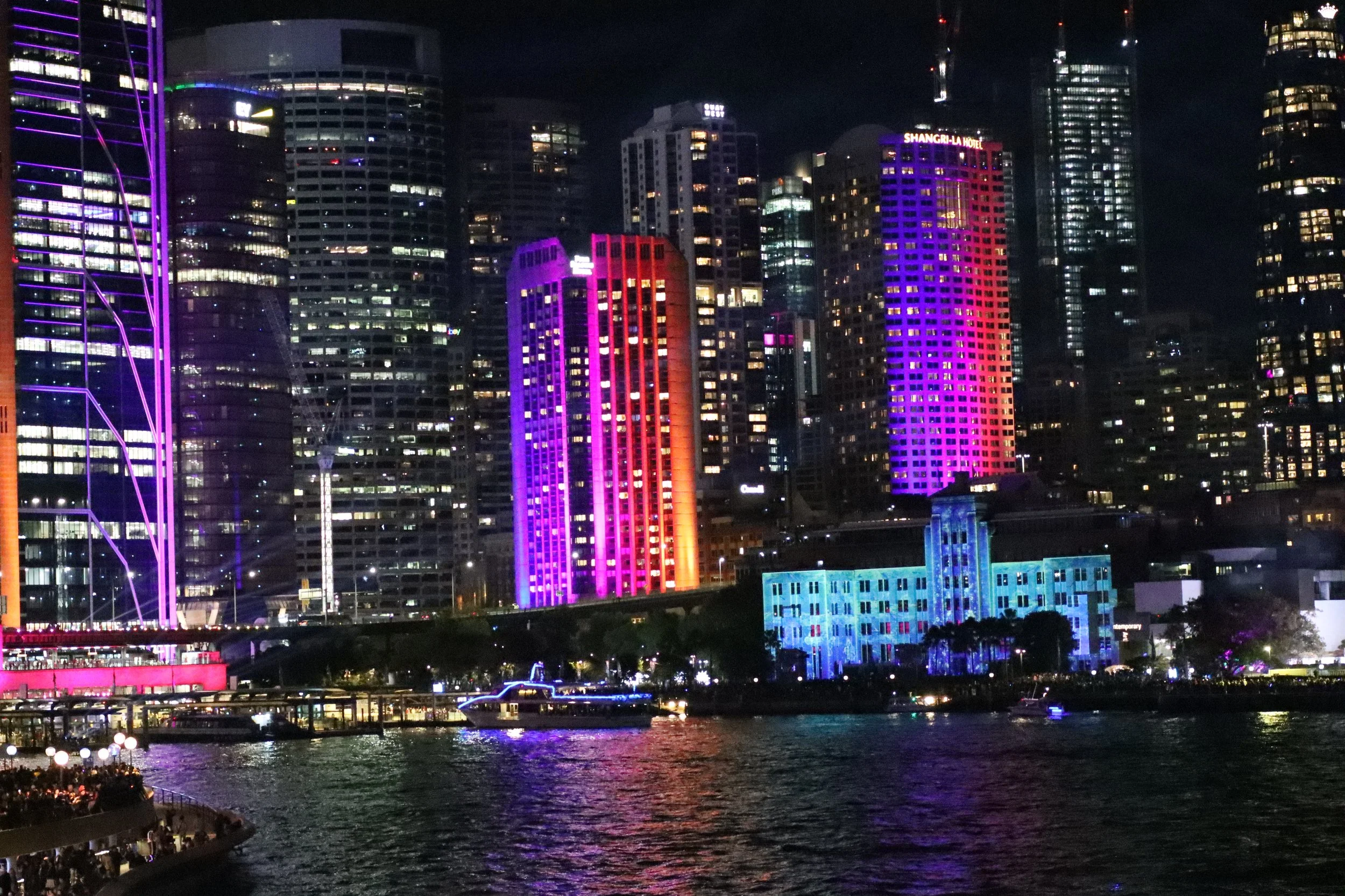 Nighttime city skyline with illuminated colorful skyscrapers reflecting on water, featuring a large purple-lit building and the Shangri-La Hotel.