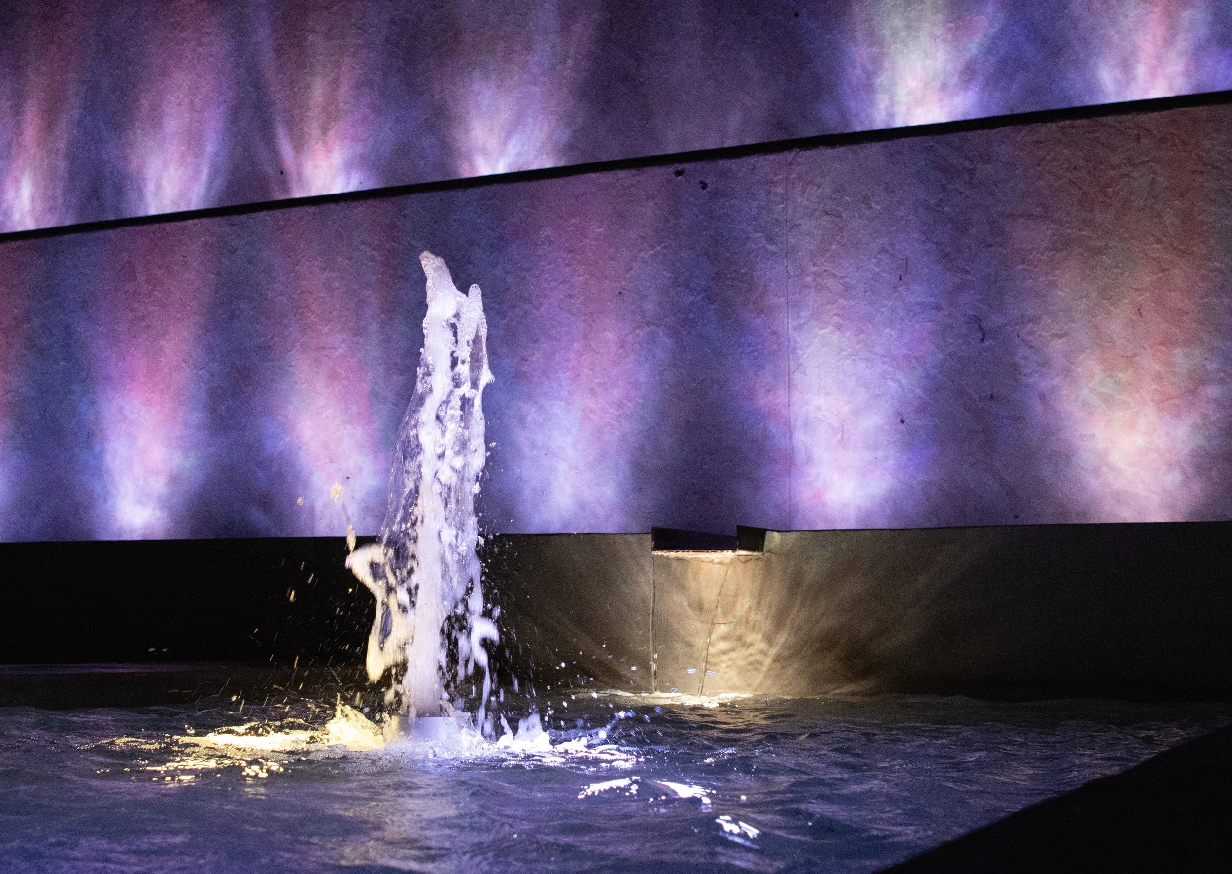 A fountain of water shooting up in front of a large, colorful, abstract background.