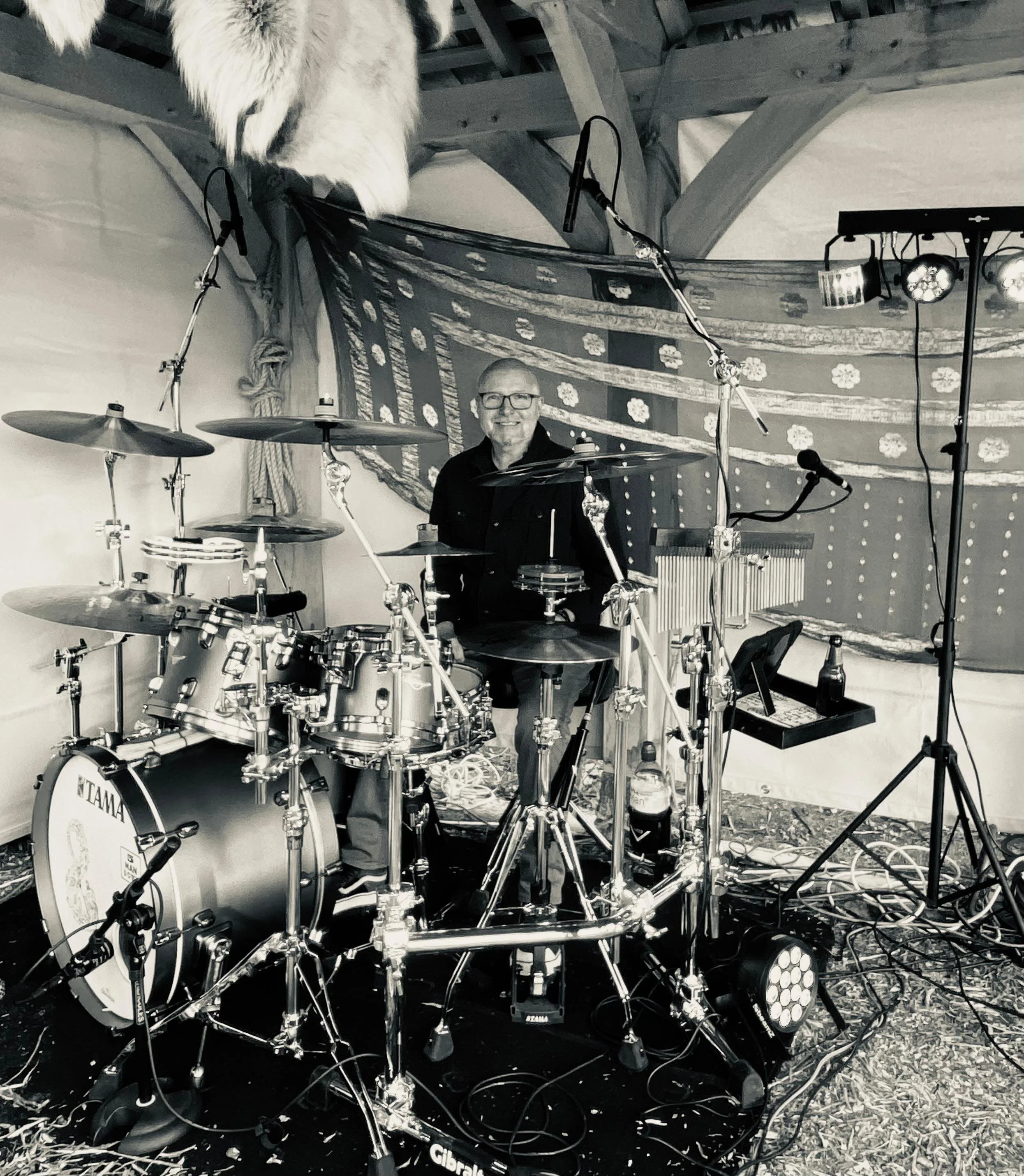 Mark Watson from 5 Man Down sitting behind a drum set, smiling, in a decorated tent with stage lights and sound equipment.
