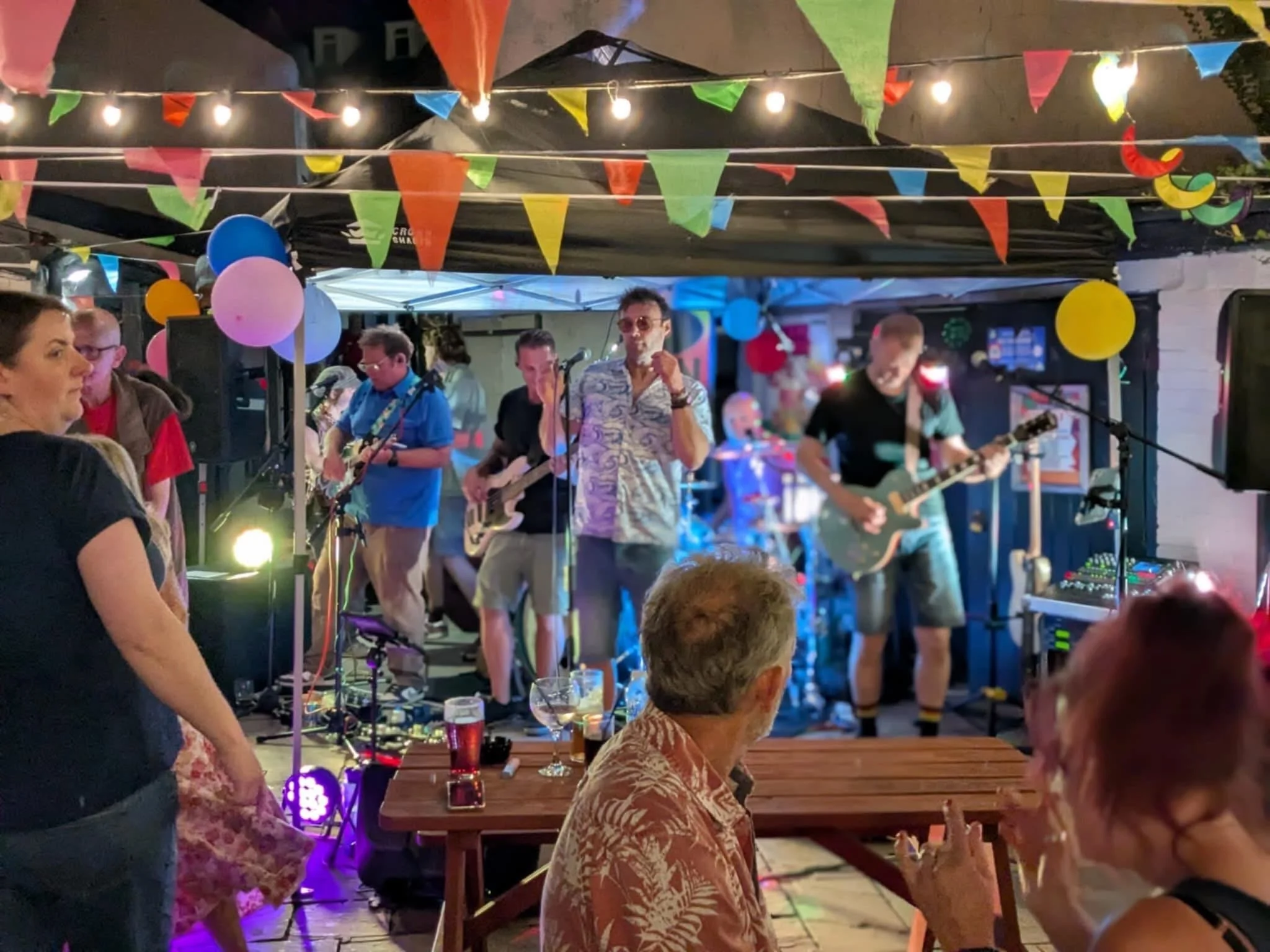 5 Man Down playing on stage at an outdoor party with colorful bunting, balloons, and string lights. People are seen enjoying the music and socializing.