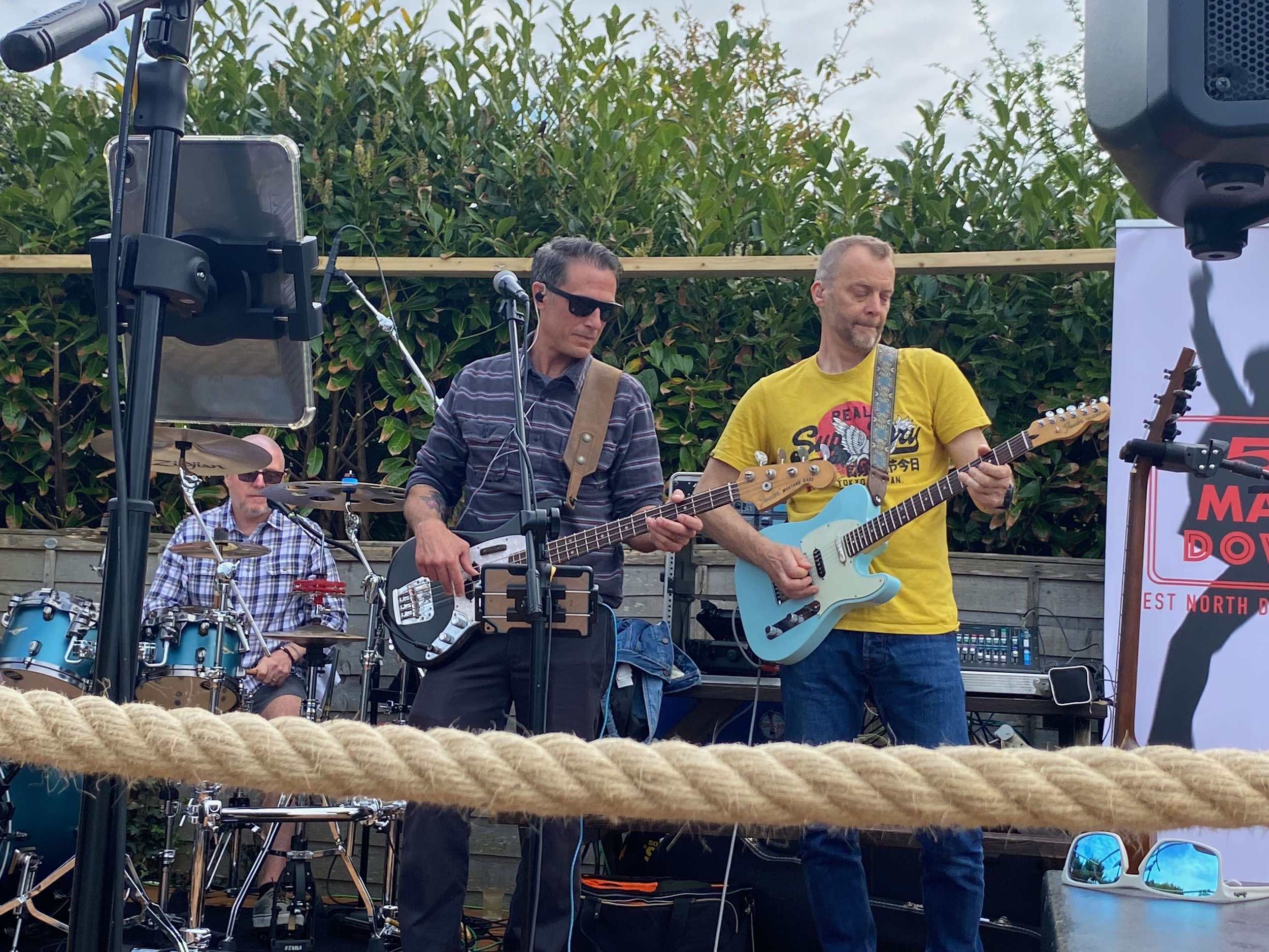 5 Man Down performing live outdoors with three members: a drummer in the background, and two guitarists in the foreground, one playing a bass guitar and the other an electric guitar, with a ladder and a banner partially visible.