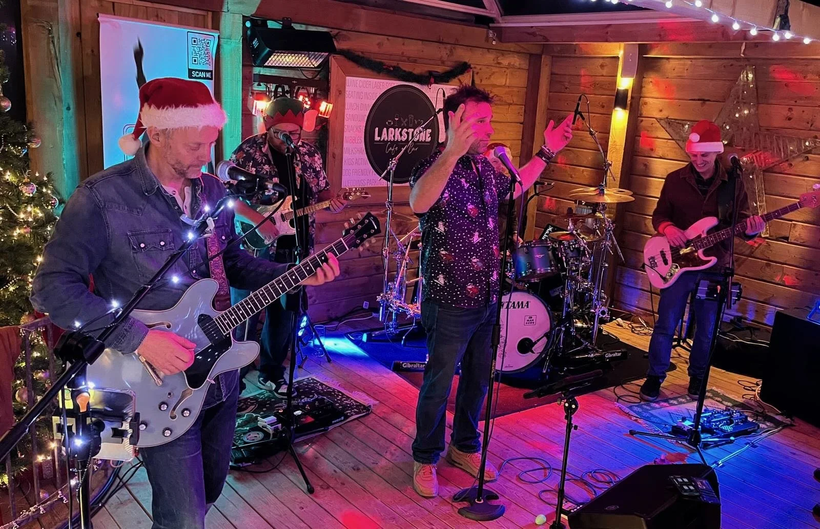 5 Man Down performing on stage with Christmas decorations, including a Christmas tree with ornaments and string lights, in a wood-paneled room. The band members are wearing Santa hats and Christmas-themed attire.