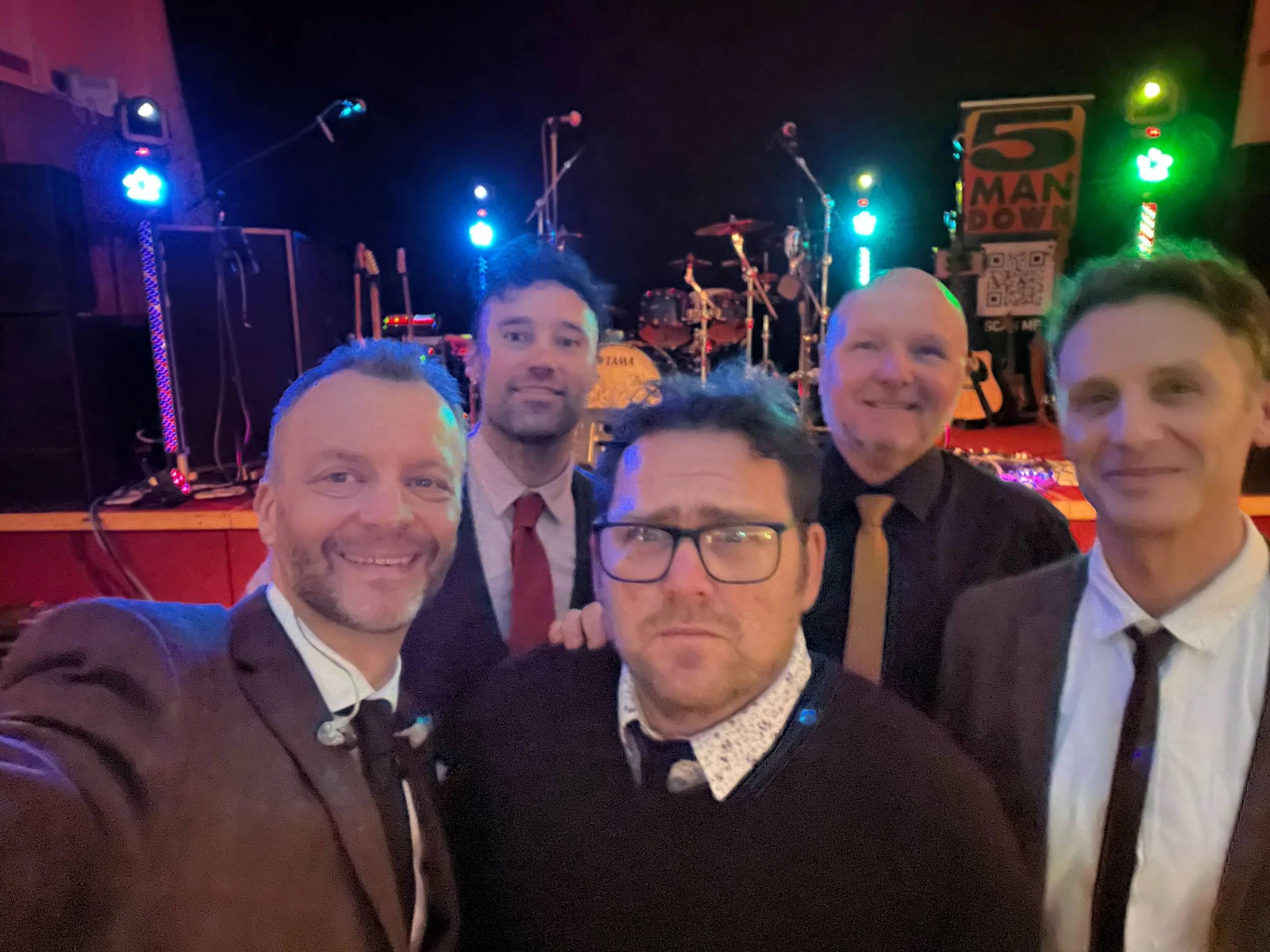 5 Man Down in formal attire taking a selfie in front of a stage with musical instruments and lighting.
