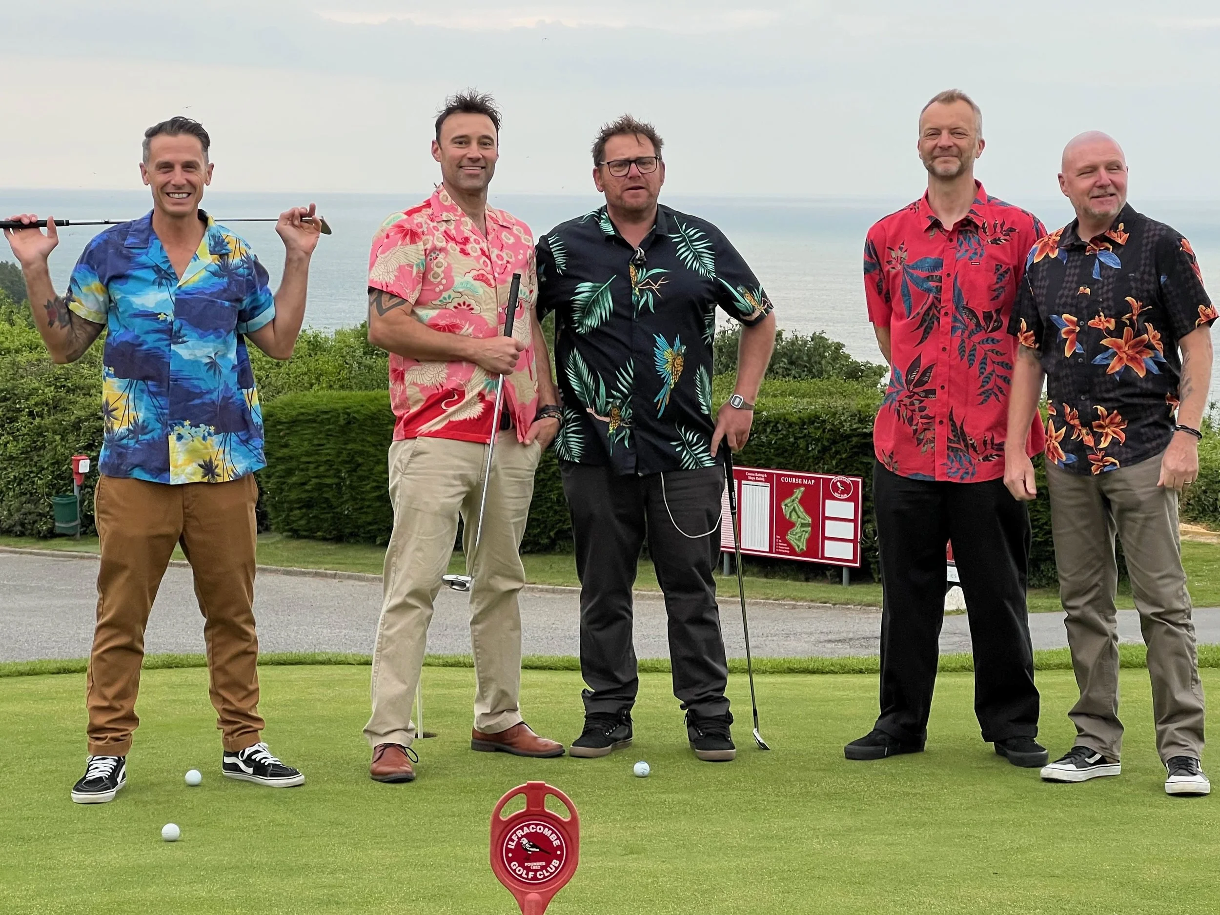 5 Man Down standing on a golf course with a view of the ocean in the background. They are wearing colorful, Hawaiian-style shirts and are holding golf clubs. There are golf balls on the ground and a course sign behind them.