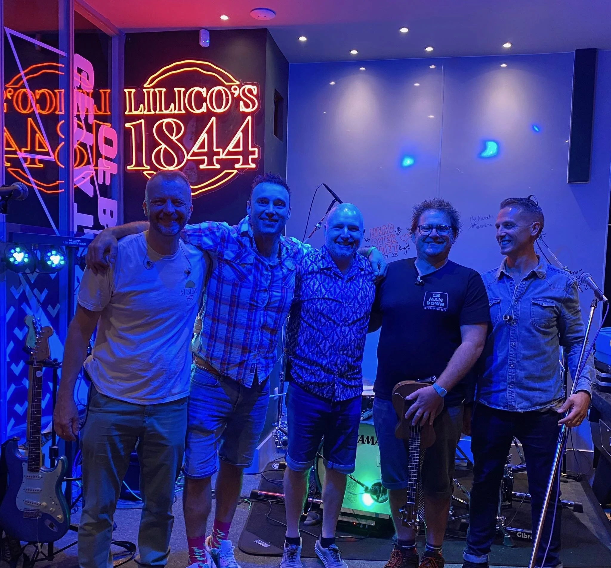 5 Man Down standing together on a stage with musical instruments, neon signs, and microphones, smiling for the photo.