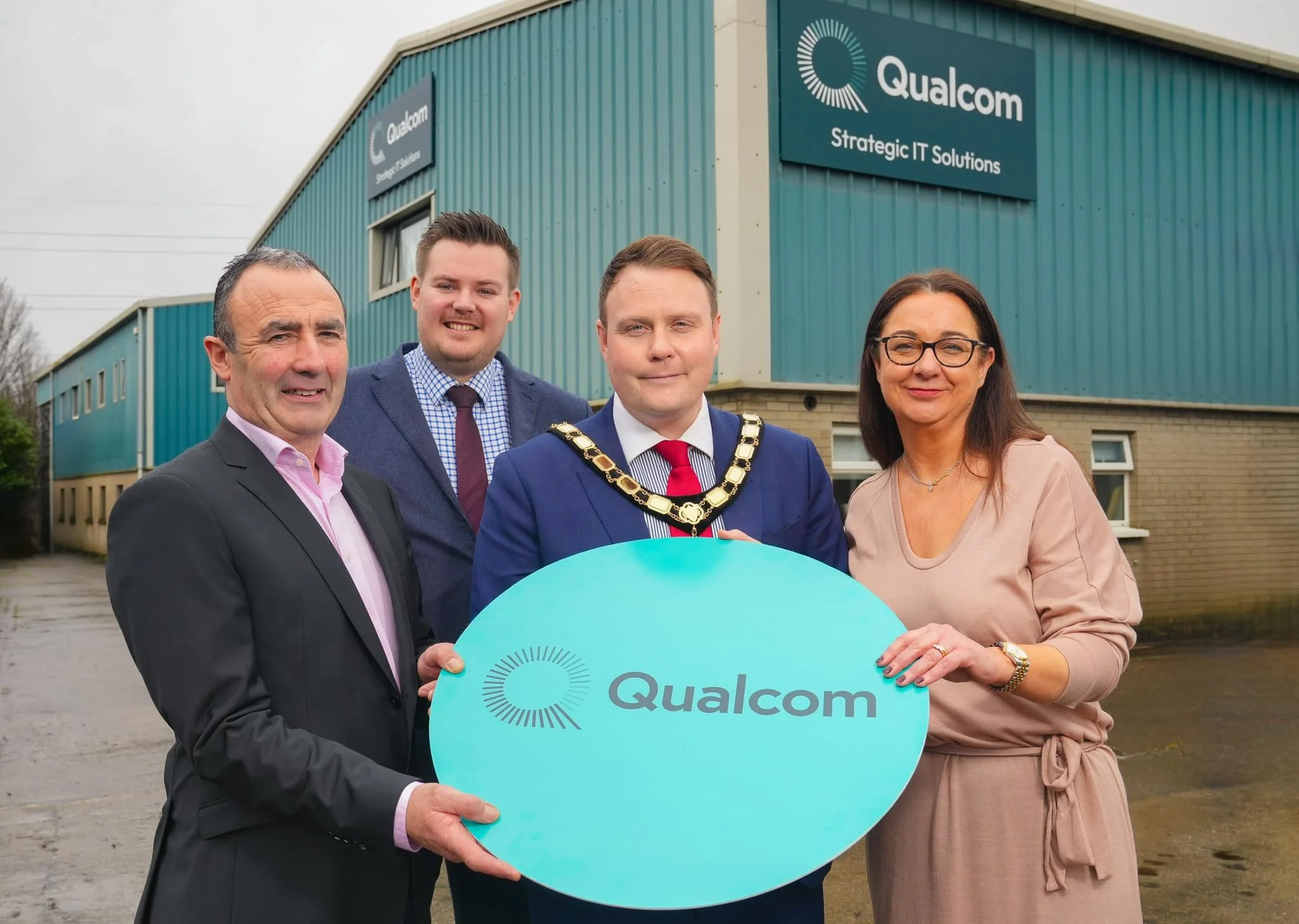 Four individuals standing outside in front of a teal building with Qualcomm signs, holding a circular Qualcomm logo sign.