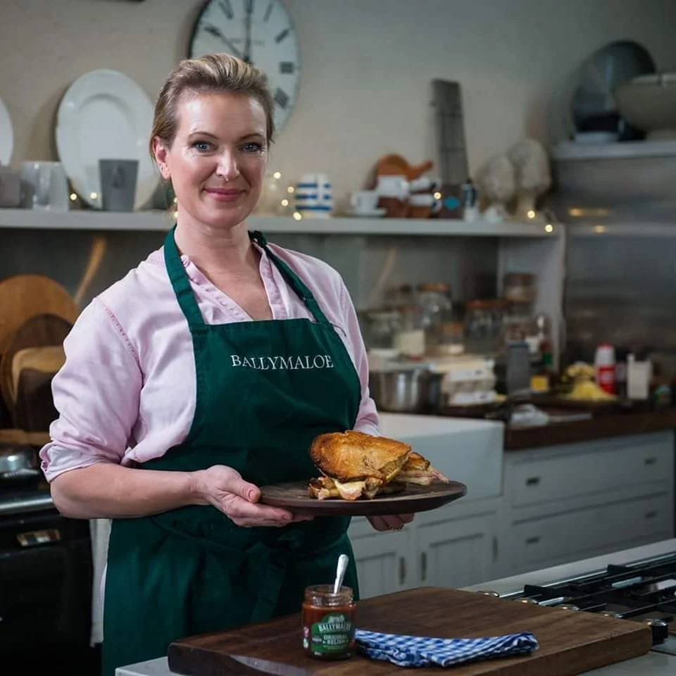 A woman wearing a green apron labeled 'Ballymaloe' stands in a kitchen, holding a plate with roasted meat. The kitchen has white cabinets, plates on shelves, and various cooking utensils and ingredients on counters.