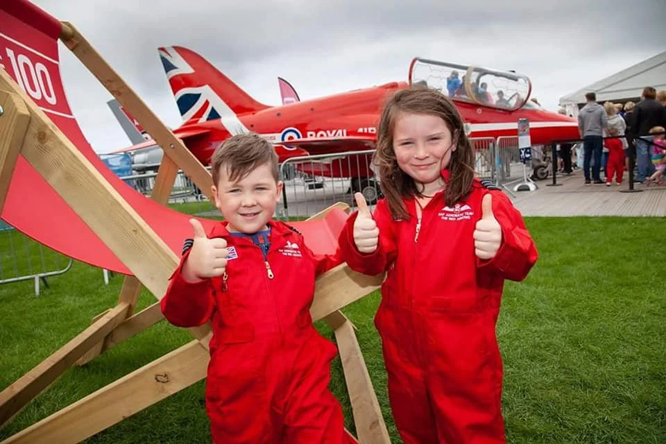 Two children in red pilot jumpsuits stand on grass and give thumbs up in front of a red aircraft and a wooden aircraft replica at an outdoor event. There are people and tents in the background.