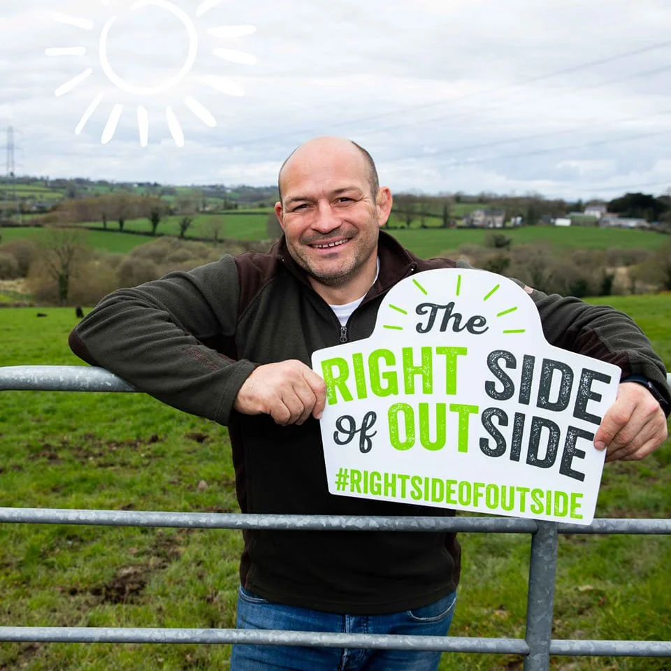 A smiling Rory Best holding a sign that says "The right side of outside" with hashtag #RIGHTSIDEOFOUTSIDE, standing outdoors near a metal gate with green fields and trees in the background, under a cloudy sky with a drawn sun in the top left corner.