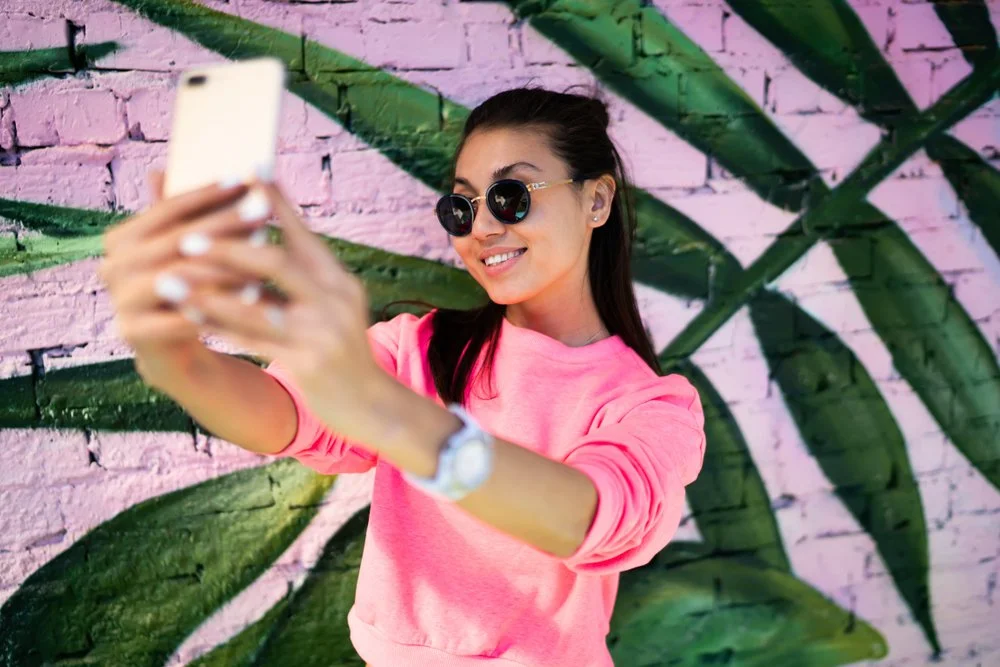 Young woman taking a selfie with a smartphone, wearing sunglasses and a pink sweatshirt, standing against a colorful graffiti wall featuring green leaves and a pink background.