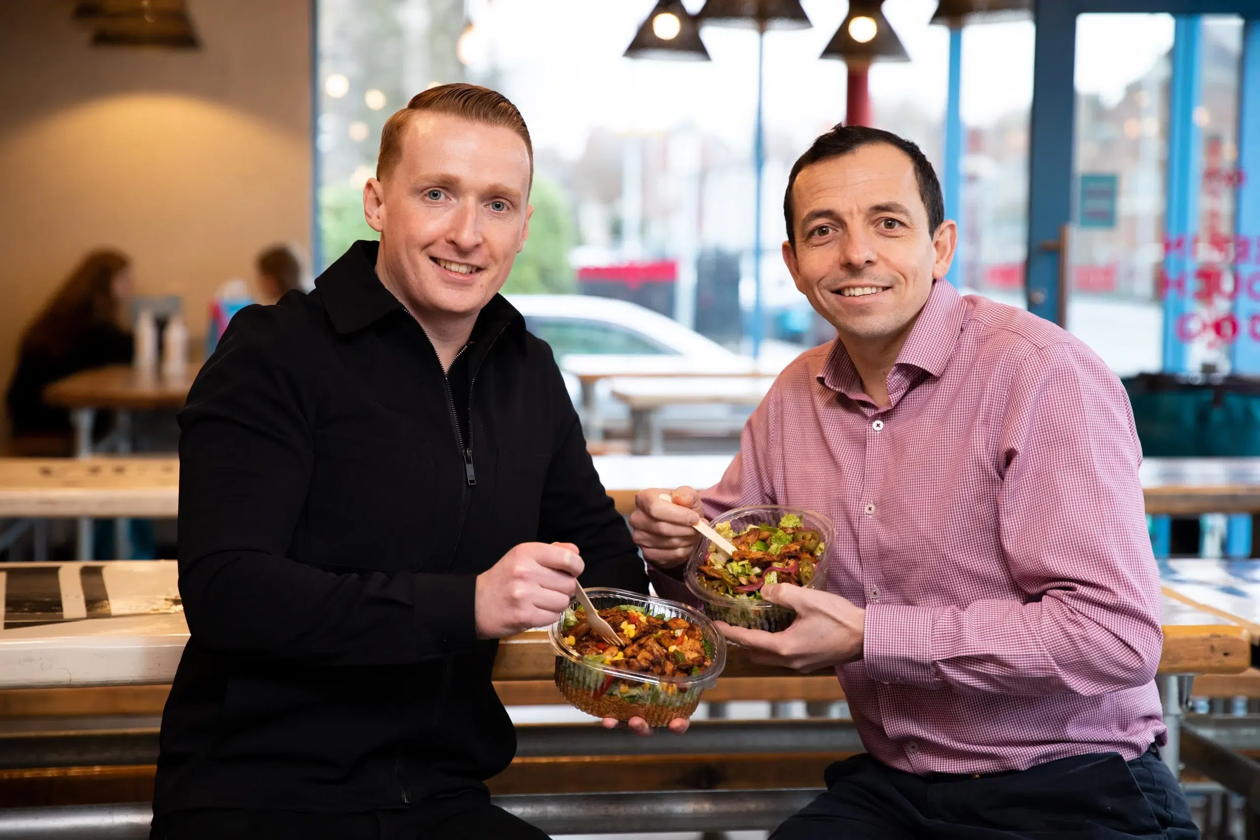 Two men sitting at a Boojum wooden table in a restaurant, smiling and holding bowls of salad. The background shows large windows with a view outside, hanging ceiling lights, and other diners.