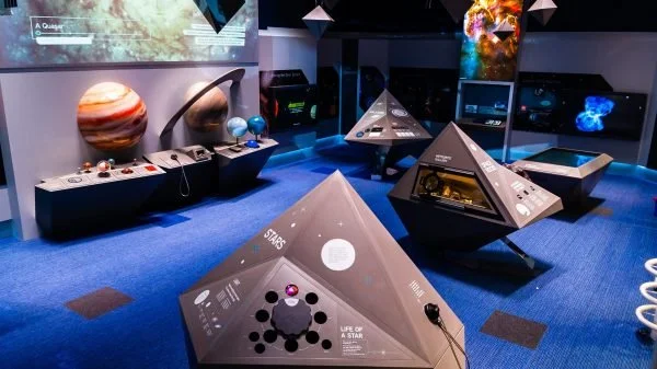Armagh Observatory and Planetarium exhibit with space-themed models, including planetary models, pyramidal displays with informational labels, and digital screens showing space imagery in a dark room with blue carpeting.